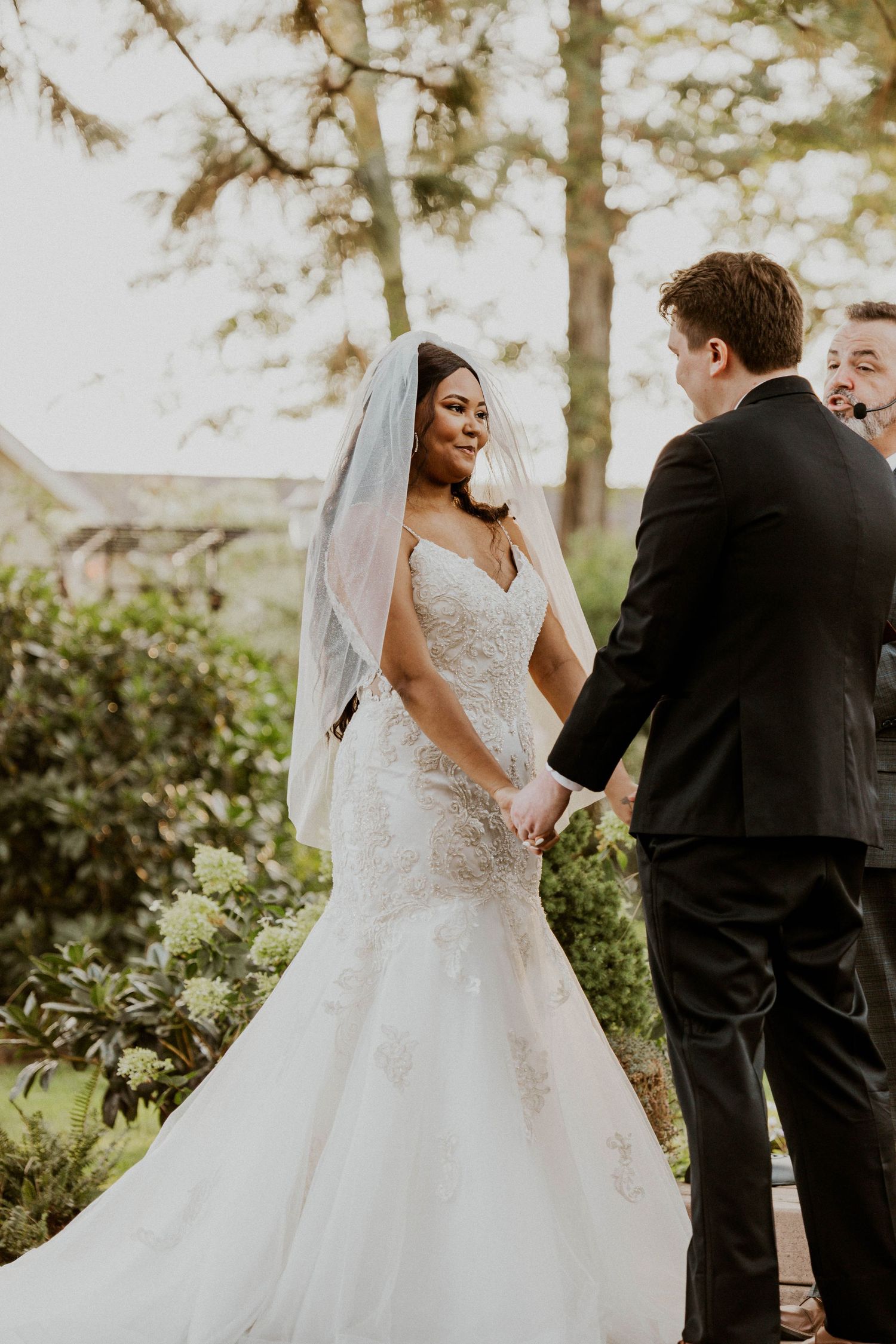 A smiling bride in a lace mermaid gown holds hands with her groom during an outdoor ceremony.