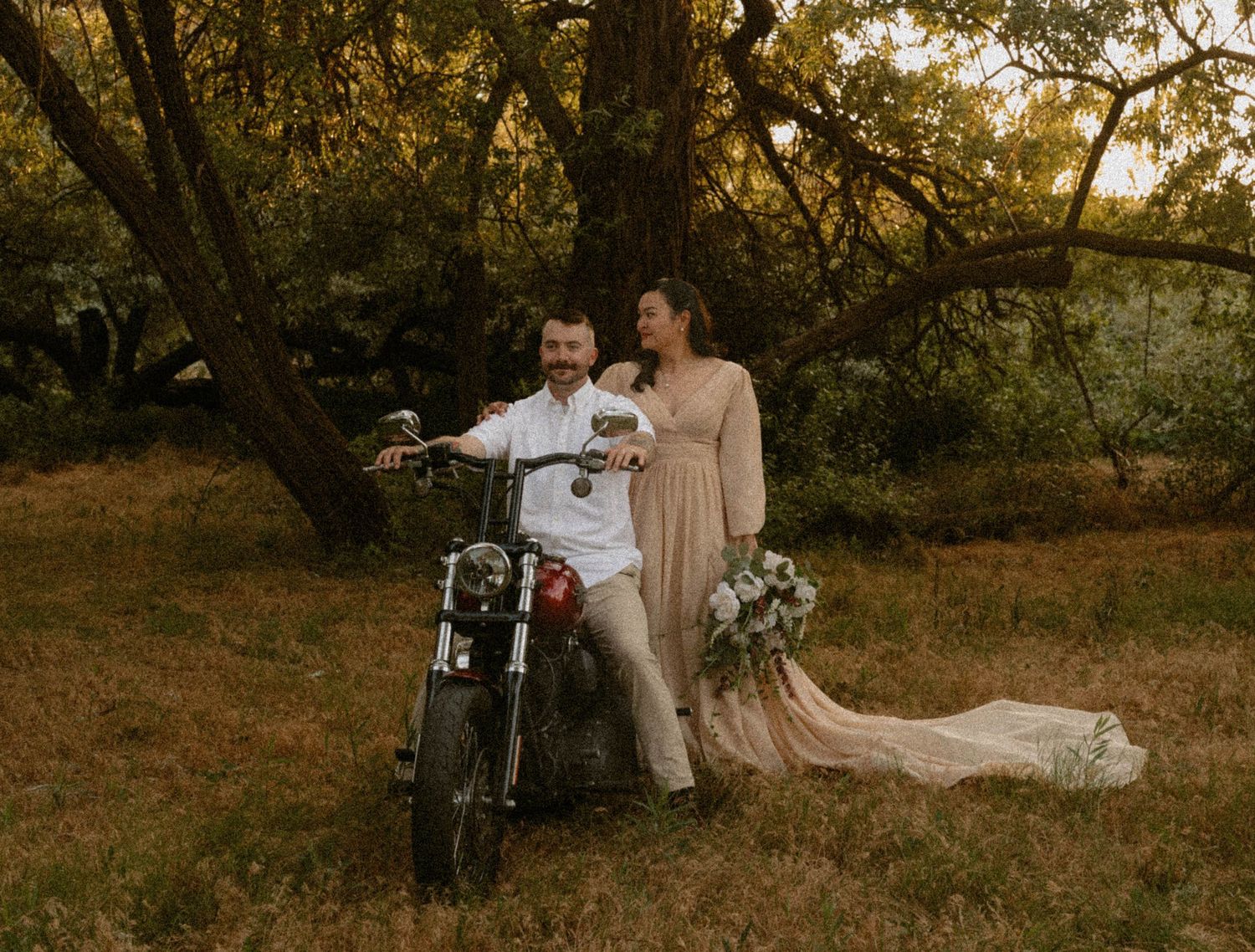 Smiling bride and groom posing beside a red motorcycle in a sunlit wooded landscape.