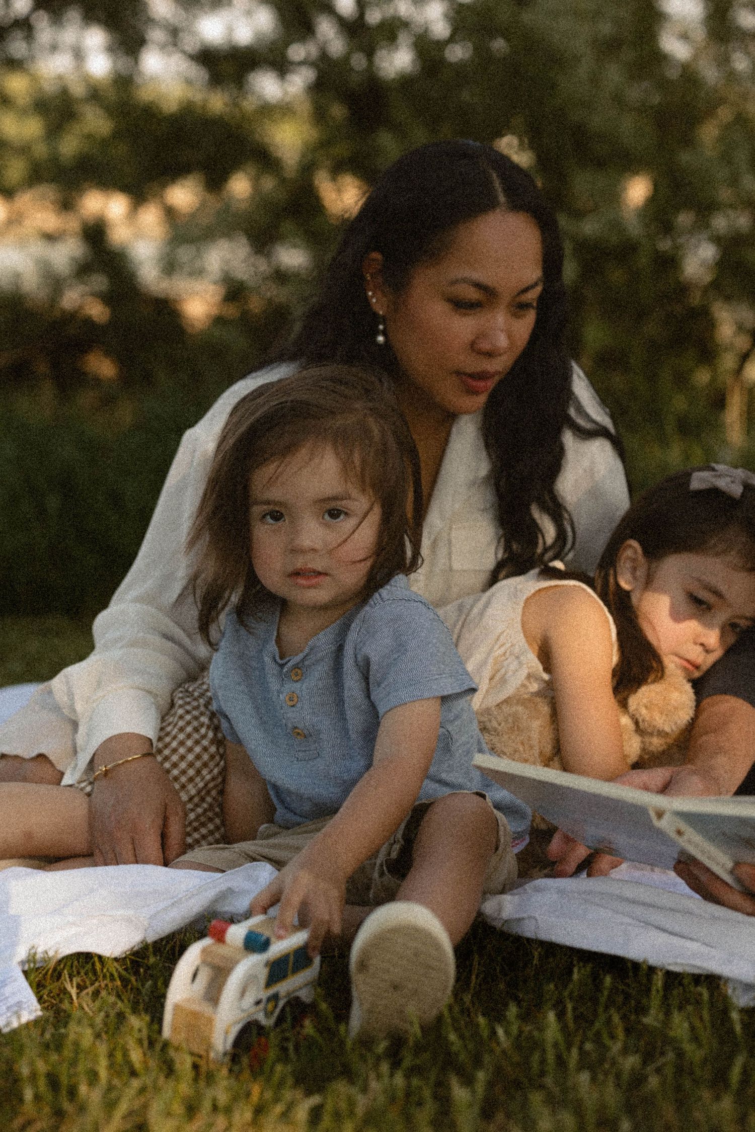 Mother sits on grass with two toddlers reading a book during a golden hour outdoor session.
