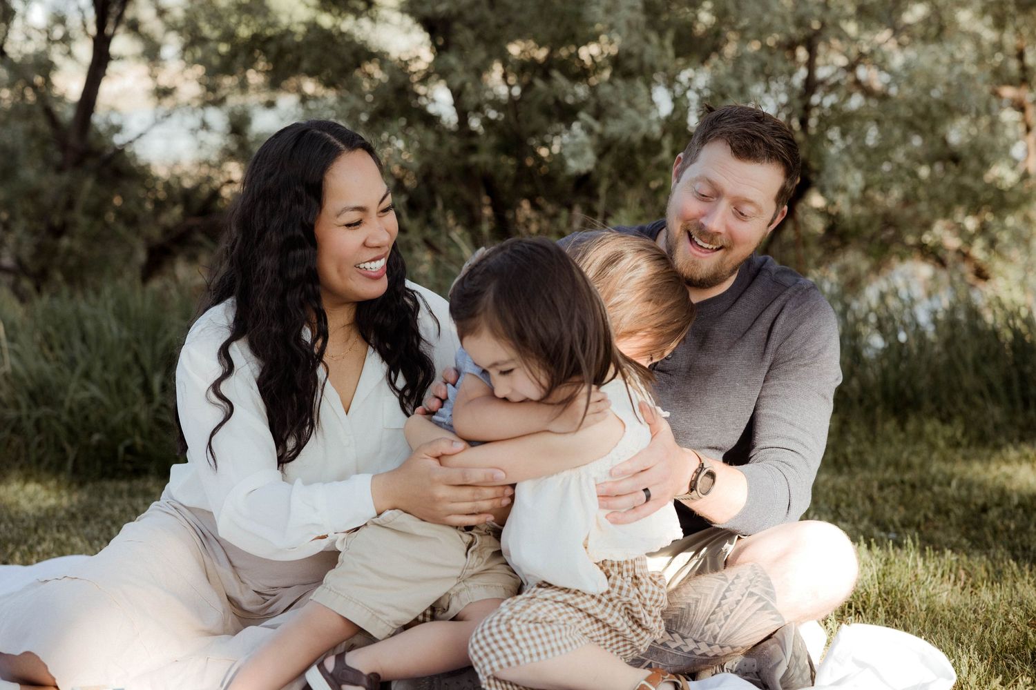 A happy family of four laughs and hugs together while sitting on a blanket in a green park.