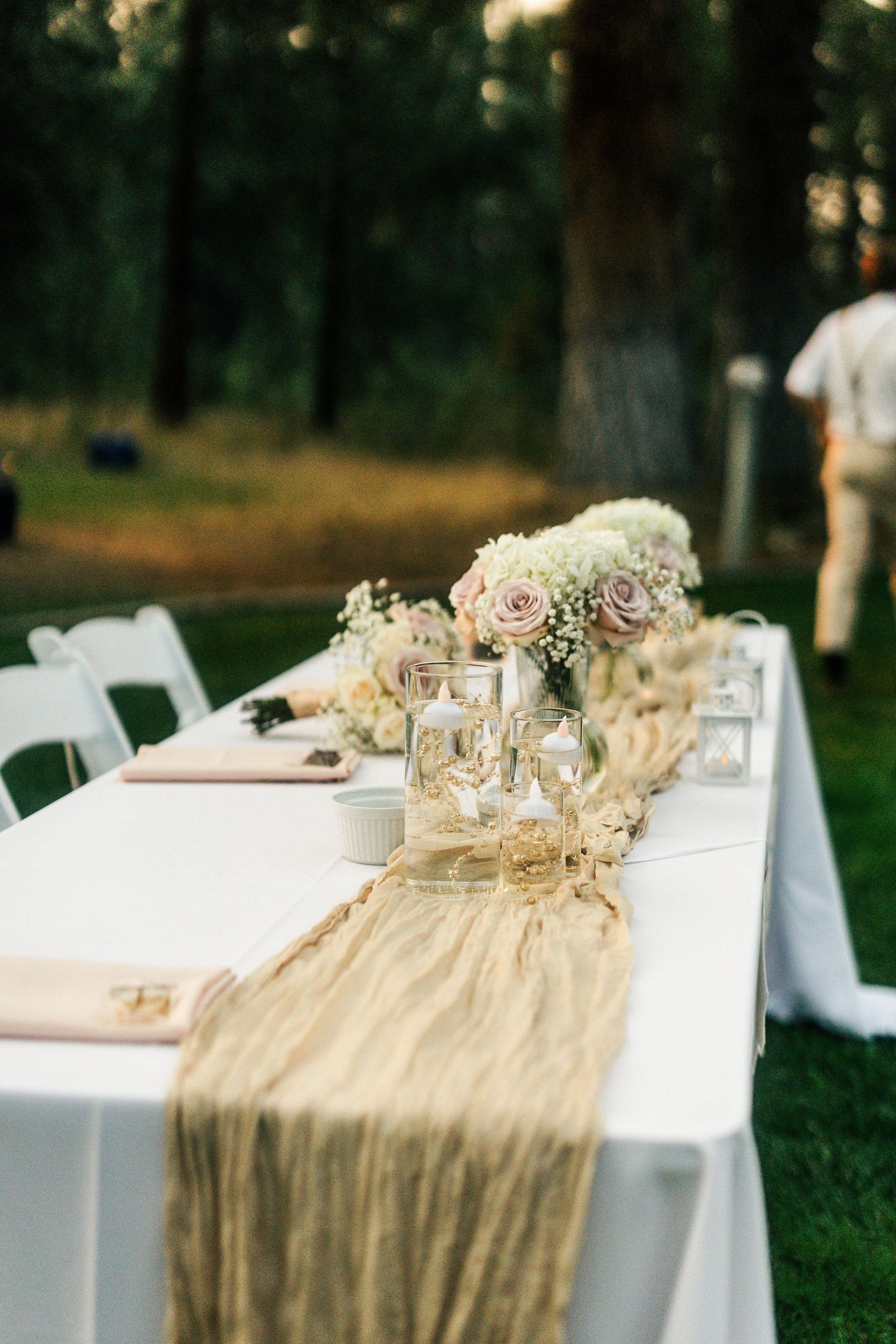 Elegant outdoor wedding reception table with burlap runner, pink roses, and candles set among tall trees.