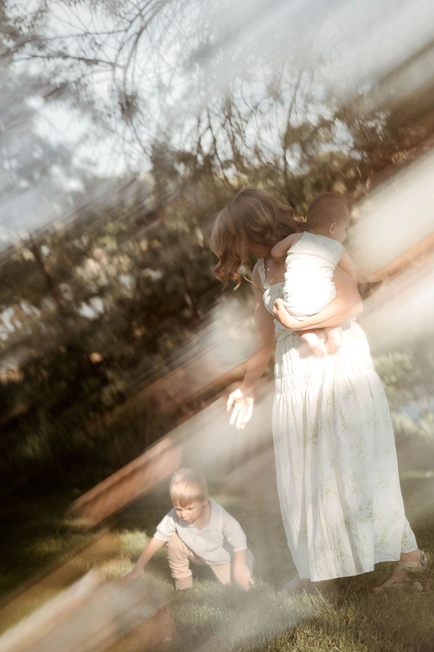 An artistic motion-blur photo of a mother in a white dress holding her baby with her toddler nearby outdoors.