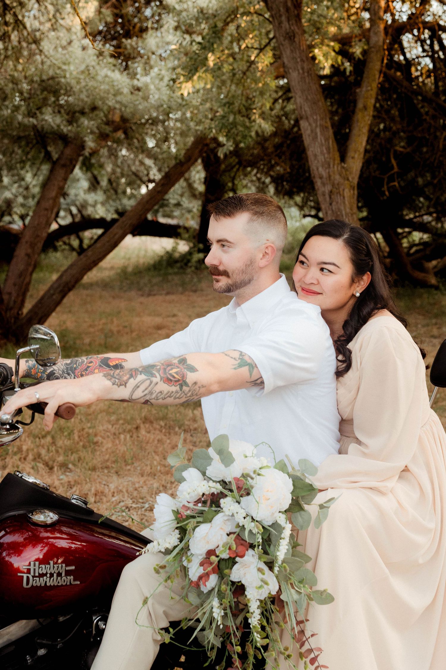Couple posing on a Harley-Davidson motorcycle outdoors, woman holding a lush white floral bouquet.