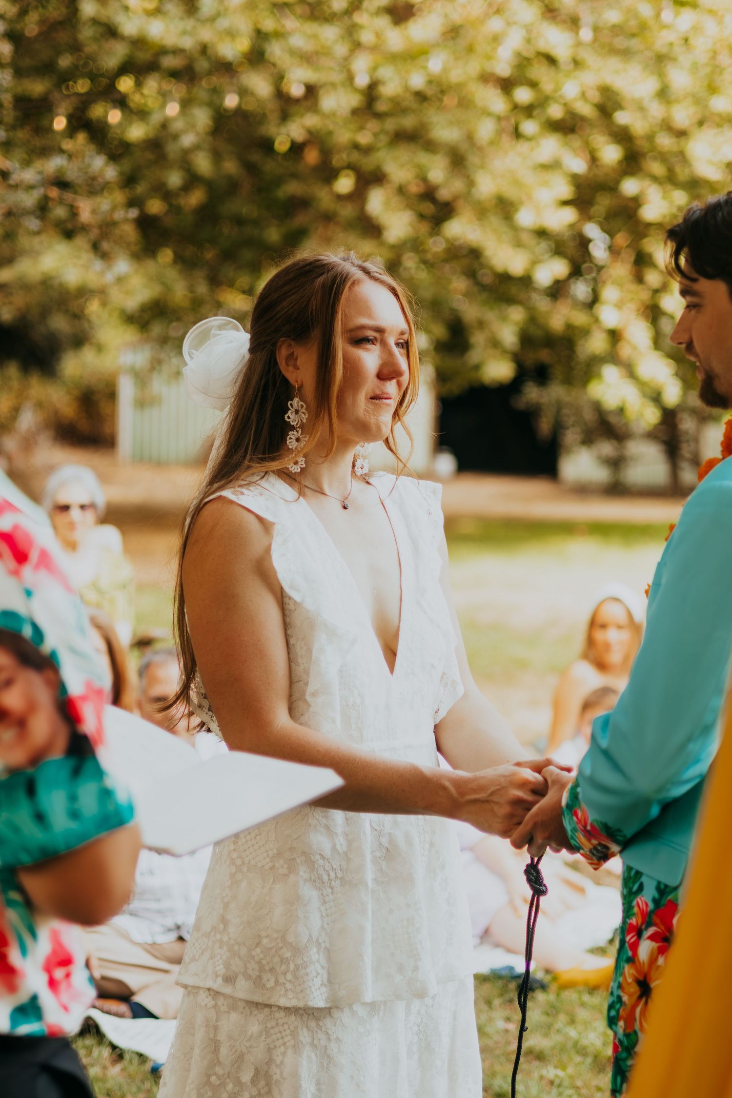 Emotional bride in a white lace tiered dress holds hands with her partner during an outdoor wedding ceremony.