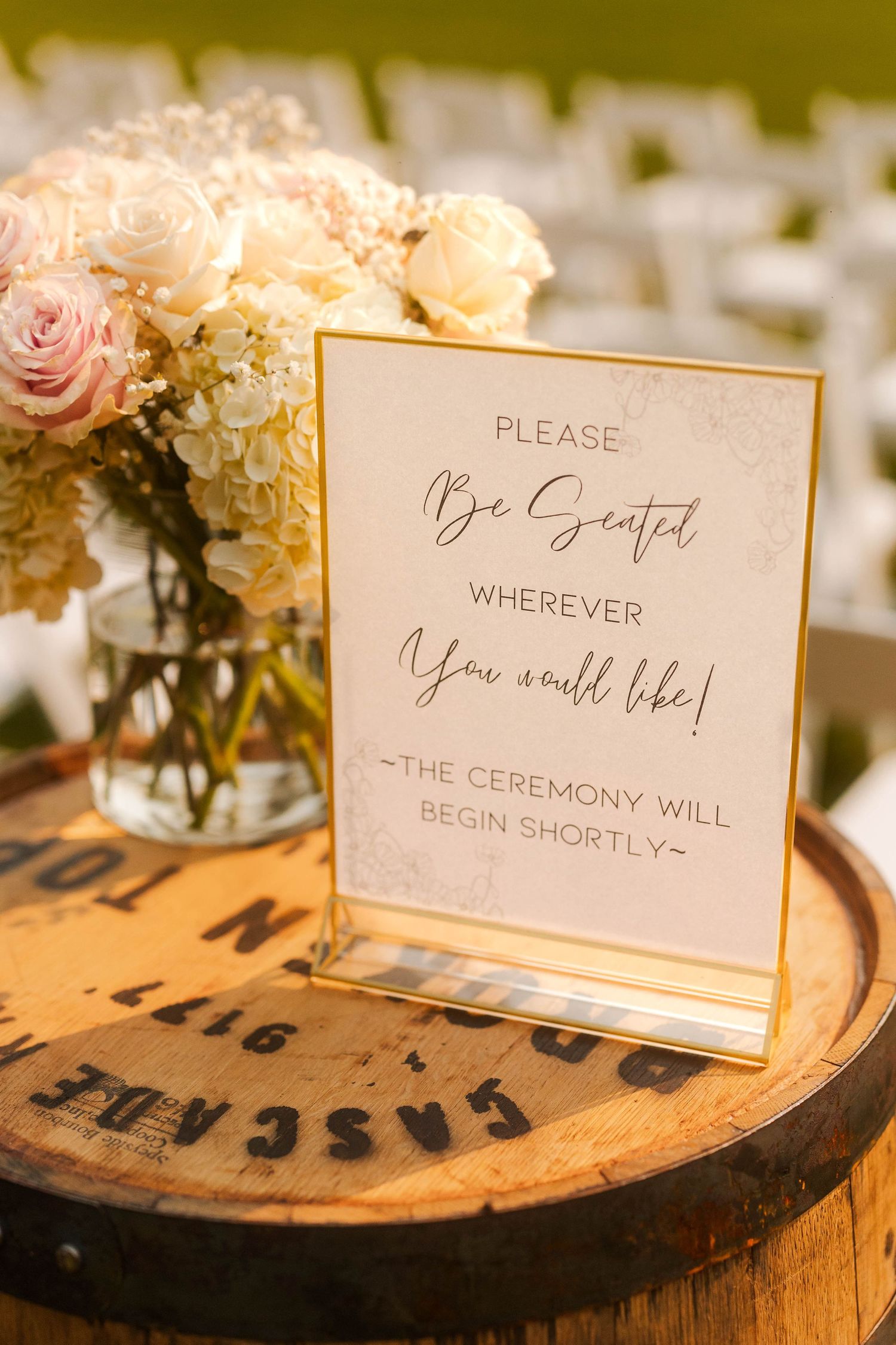 Wedding ceremony sign on barrel reading please be seated wherever you would like beside floral arrangement.