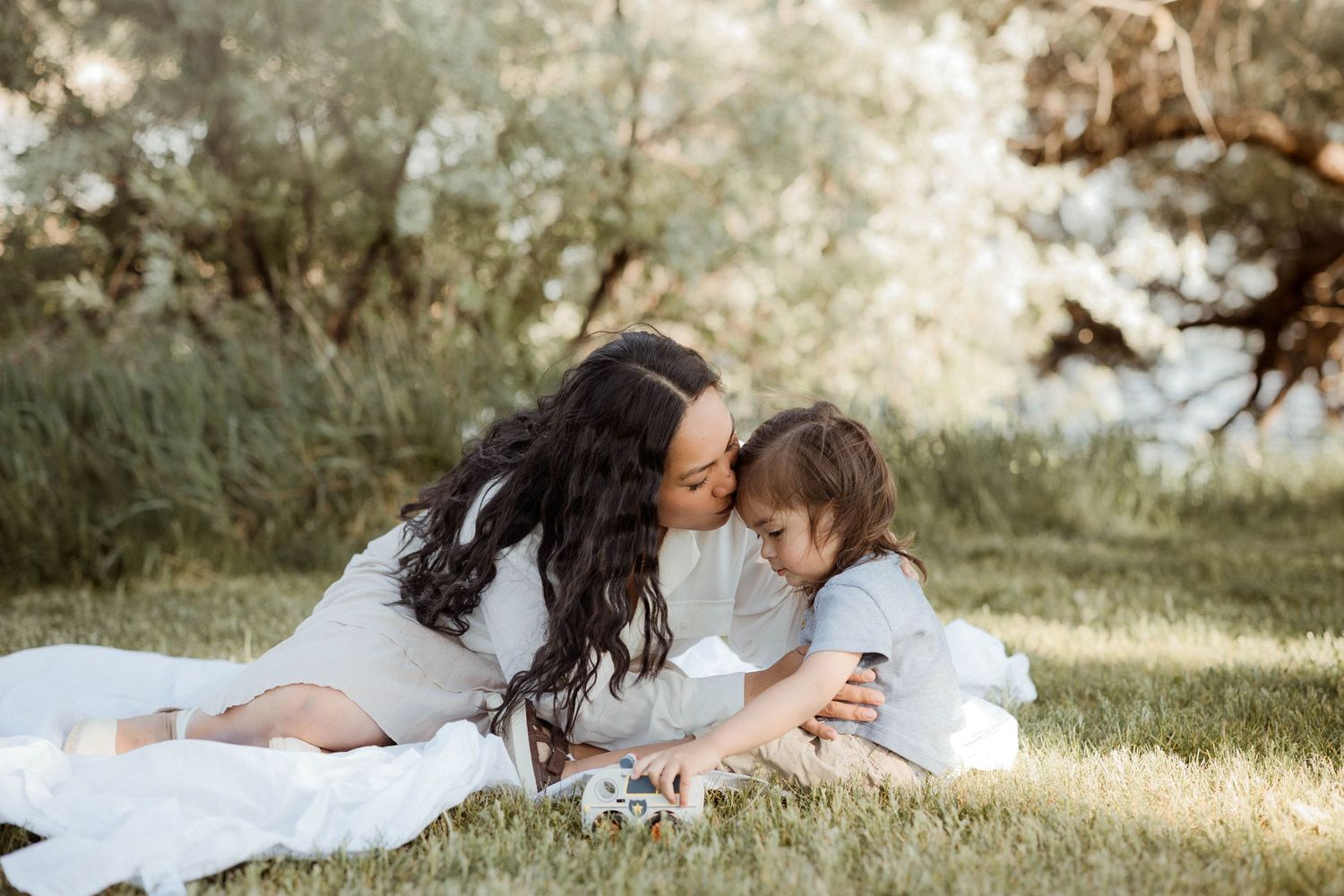 A mother tenderly kisses her young child while lying on a white blanket in a lush green park.