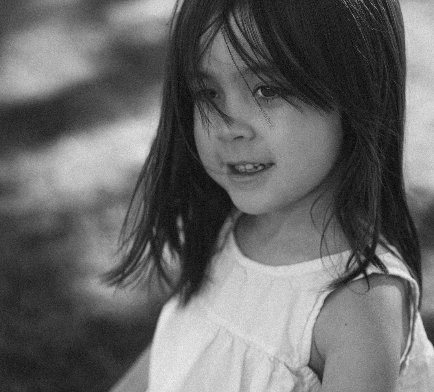 A black and white portrait of a young girl with dark hair wearing a white sleeveless dress outdoors.
