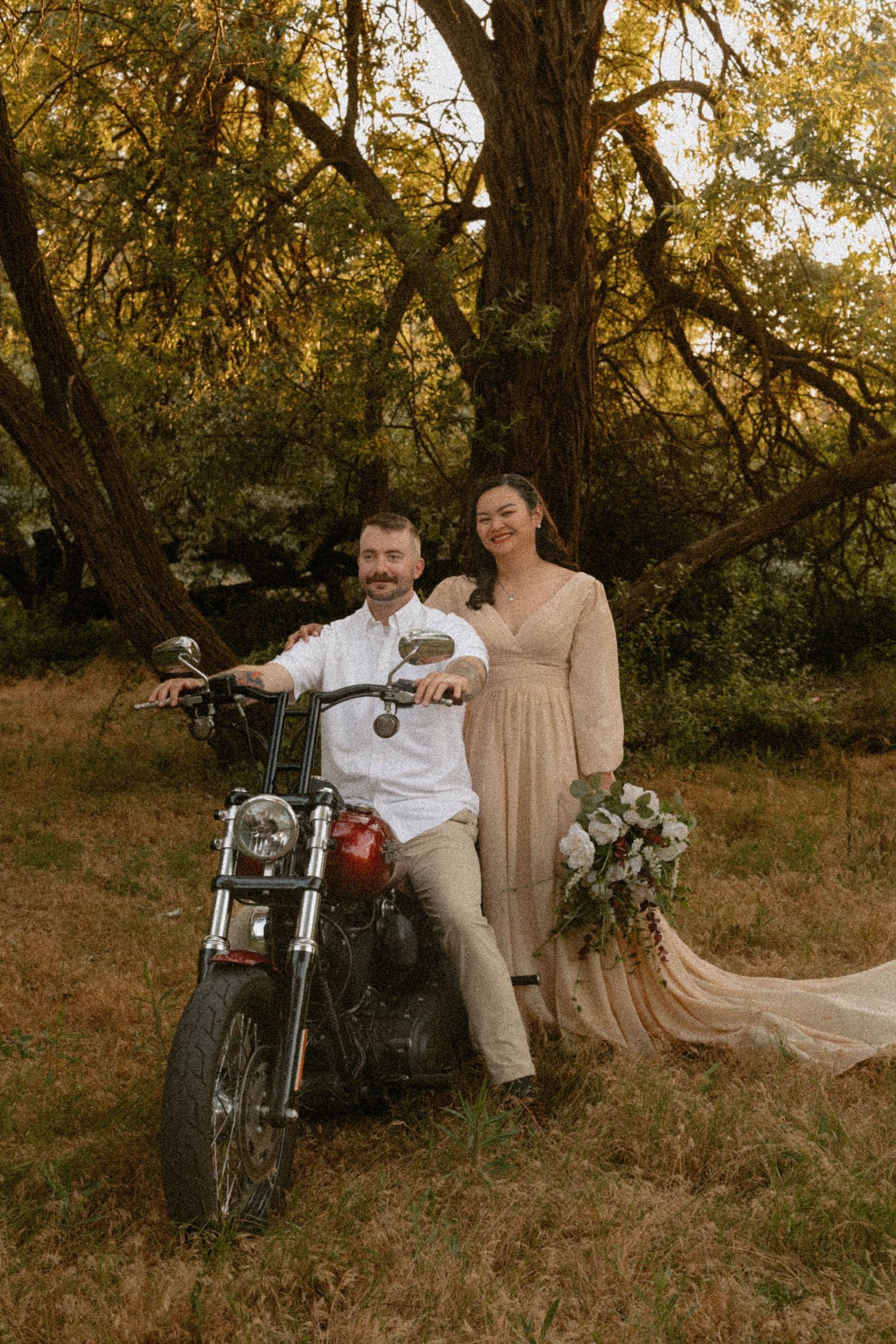 Bride and groom posing with a red motorcycle in a wooded outdoor setting at golden hour.