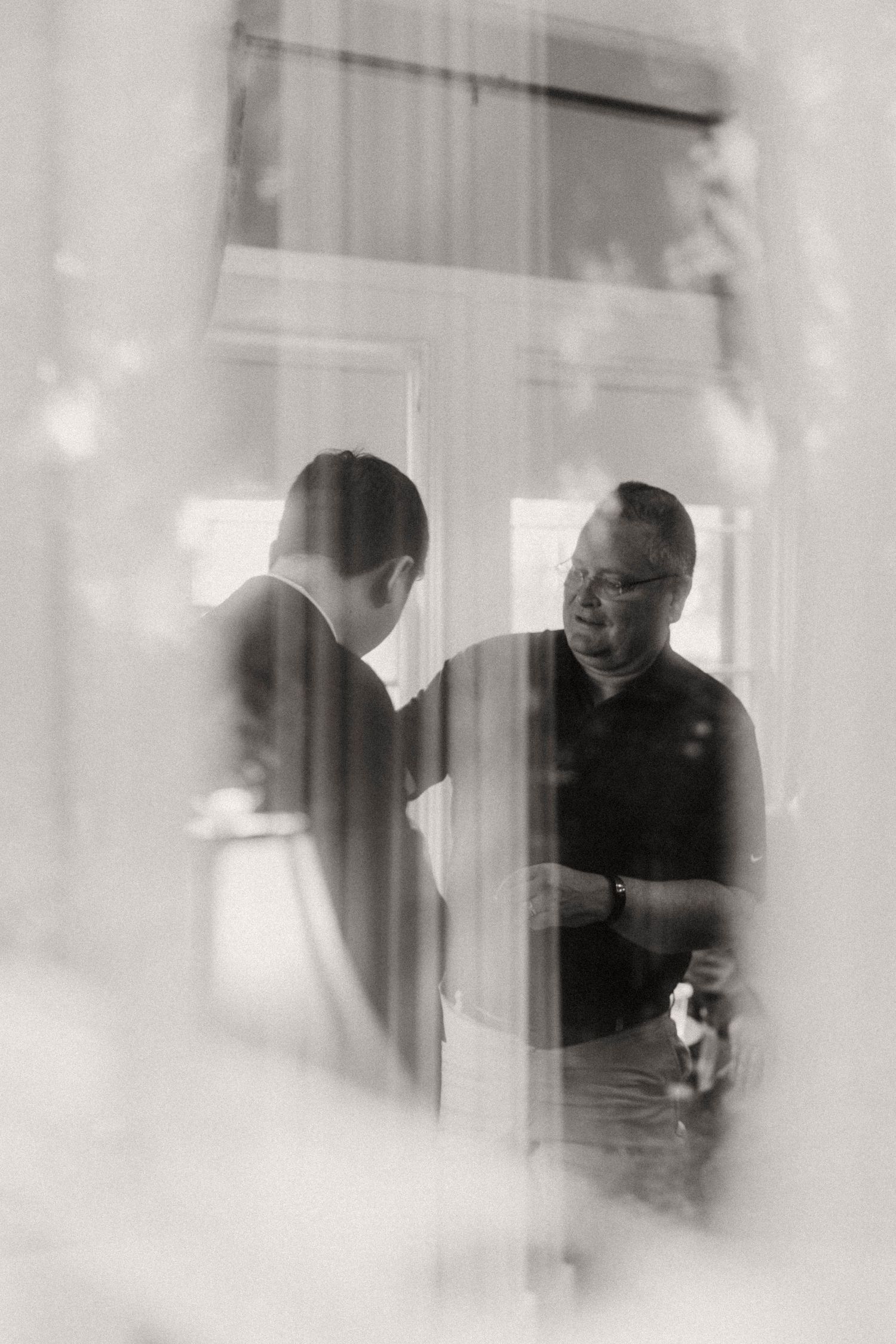 Black and white candid photo of two men conversing near a window before a wedding ceremony.