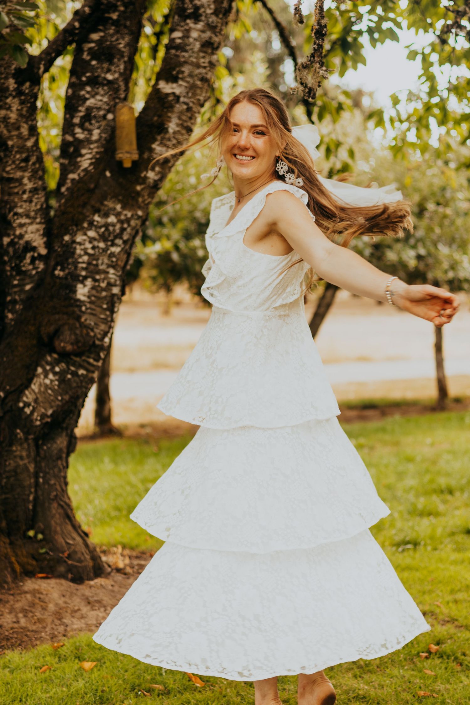 Joyful bride in a tiered white lace dress twirls and dances outdoors near a tree on her wedding day.