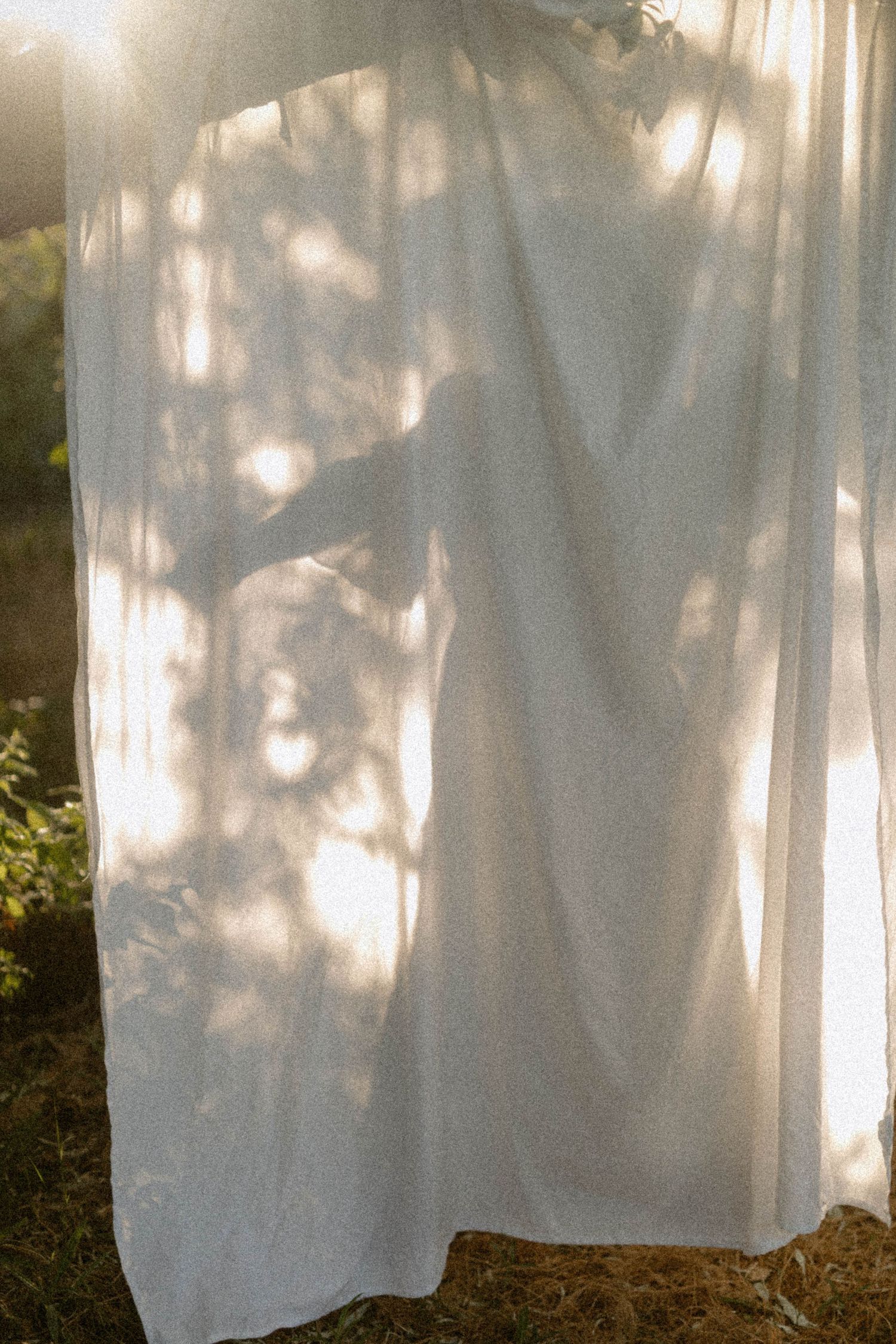 White sheer curtain hanging outdoors with sunlight filtering through, casting tree shadows on the fabric.