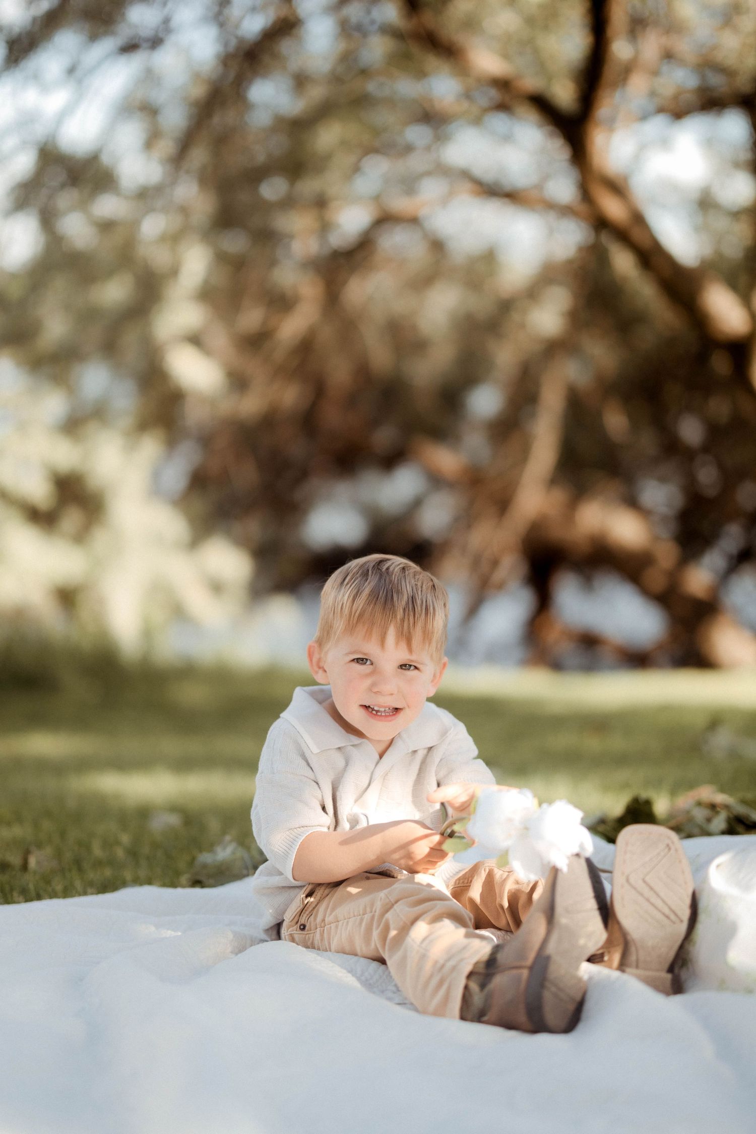 A smiling blonde toddler boy in a cream outfit sits on a white blanket on green grass in warm sunlight.