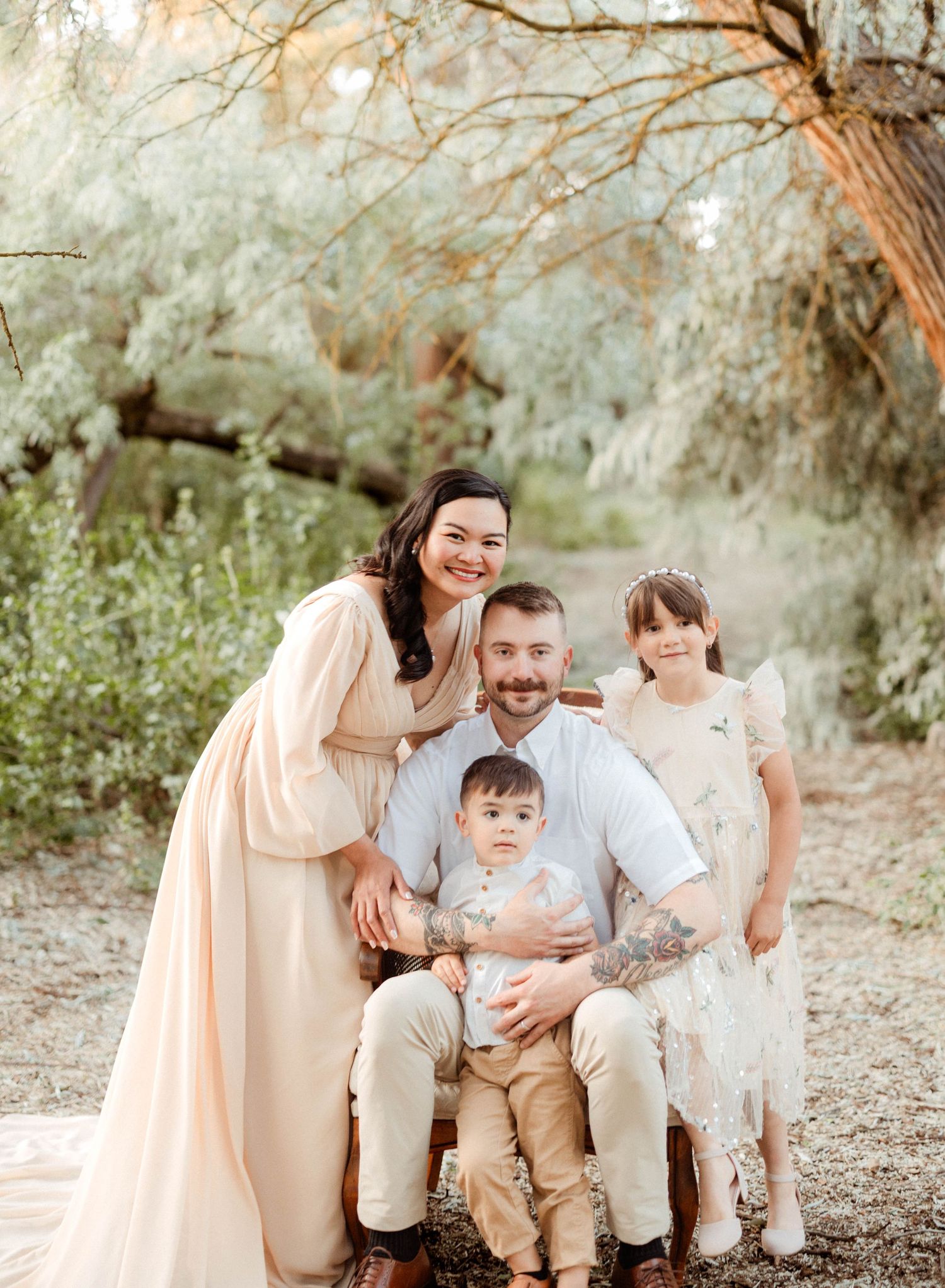 A smiling family of four poses outdoors under soft green trees in neutral tones for a professional portrait session.