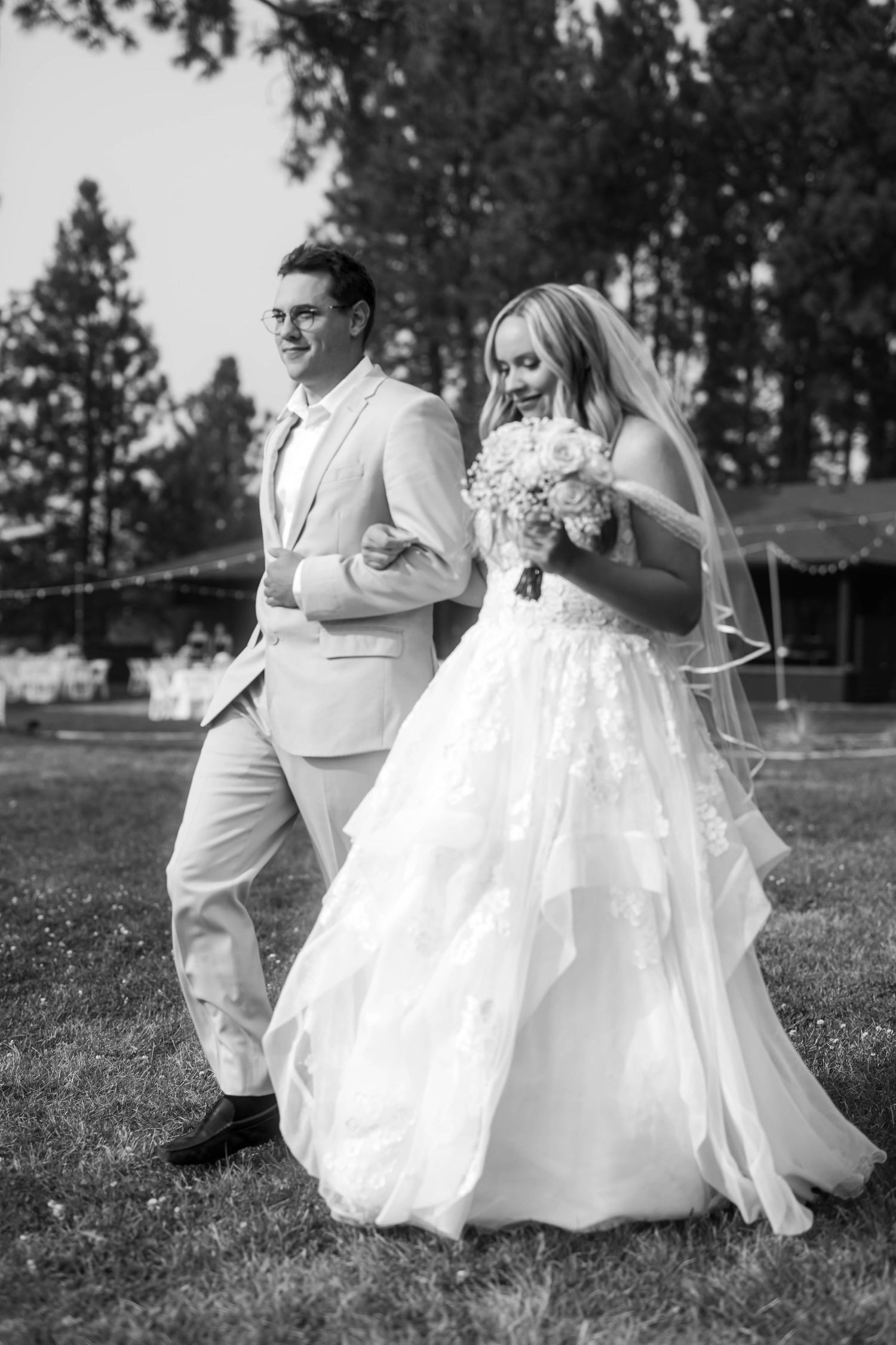Black and white photo of a smiling groom in a tan suit walking with his bride in a flowing white gown.