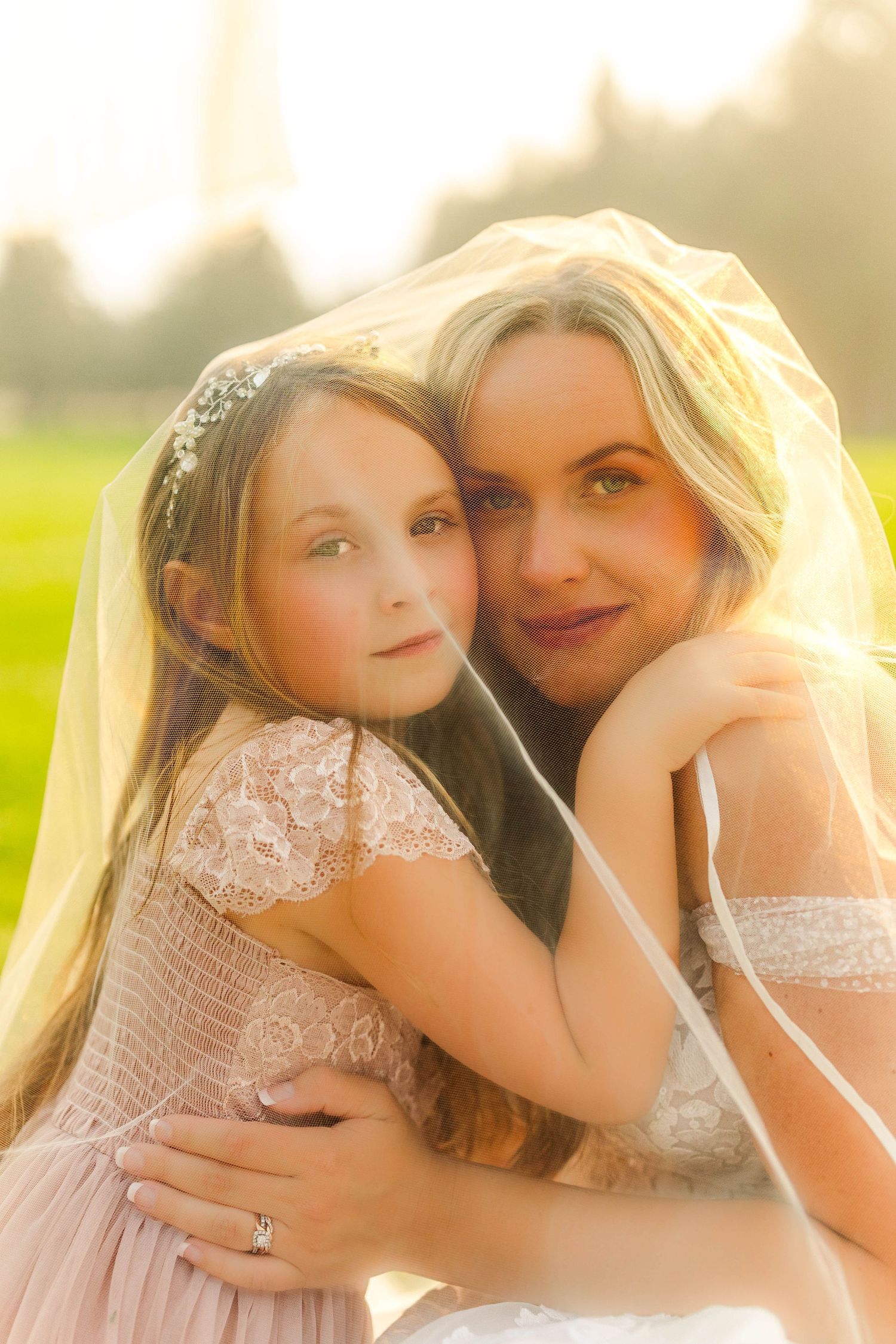 Bride in white veil and flower girl in pink lace dress embracing outdoors in golden sunlight.