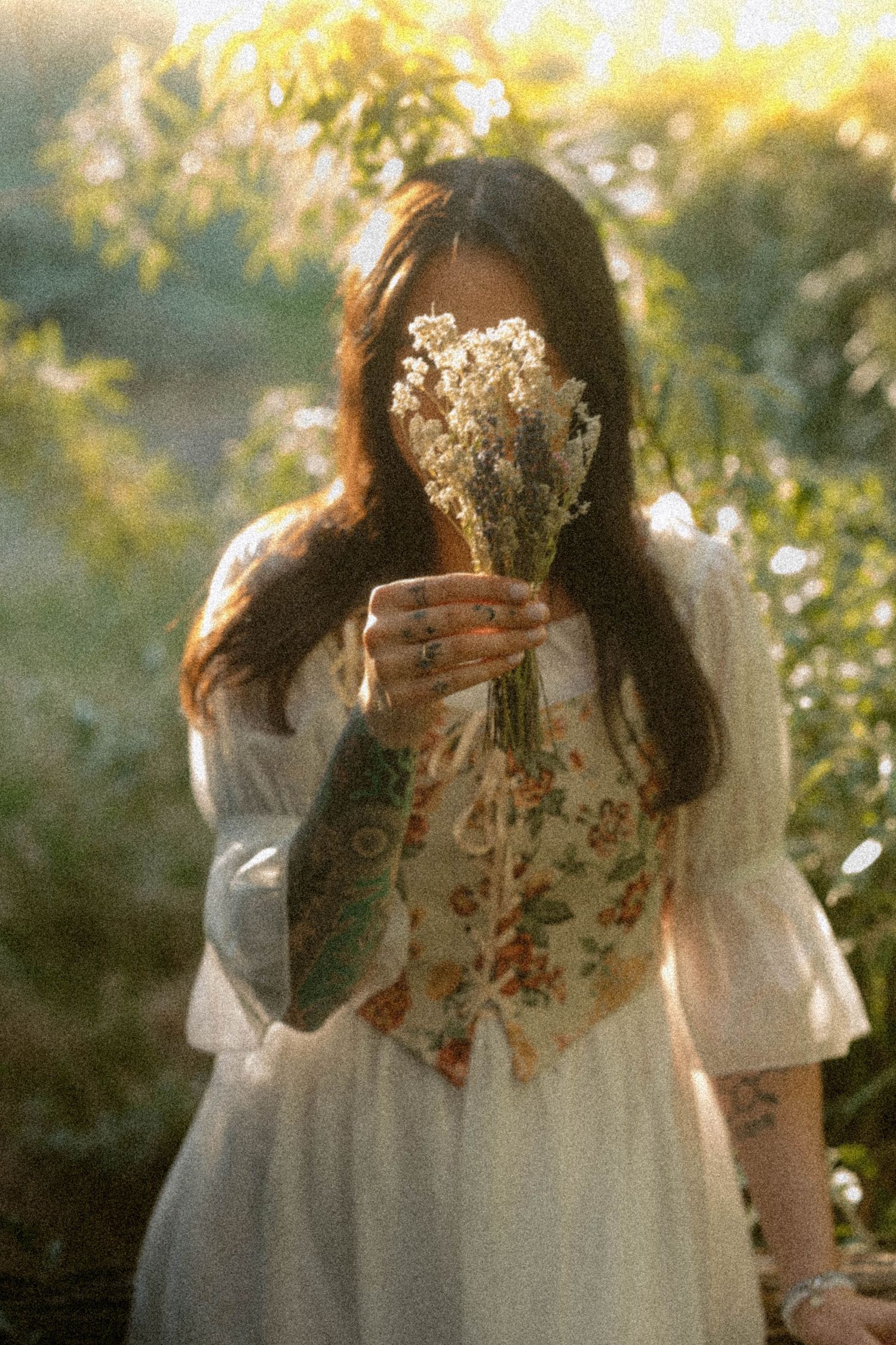 Woman in white dress and floral corset holds wildflower bouquet in golden sunlight among lush greenery.