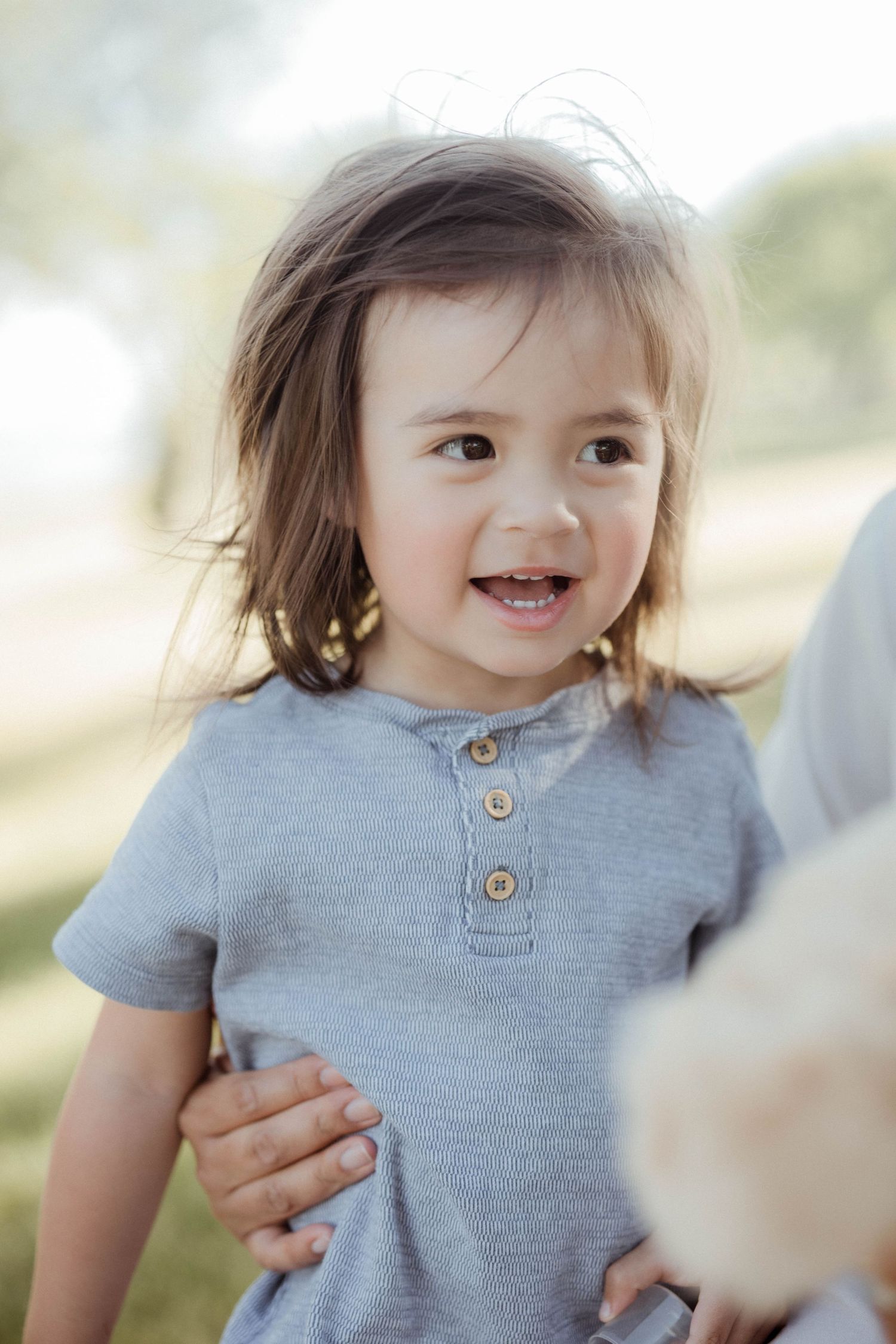 Happy toddler with brown hair in gray romper smiling in bright outdoor park setting.