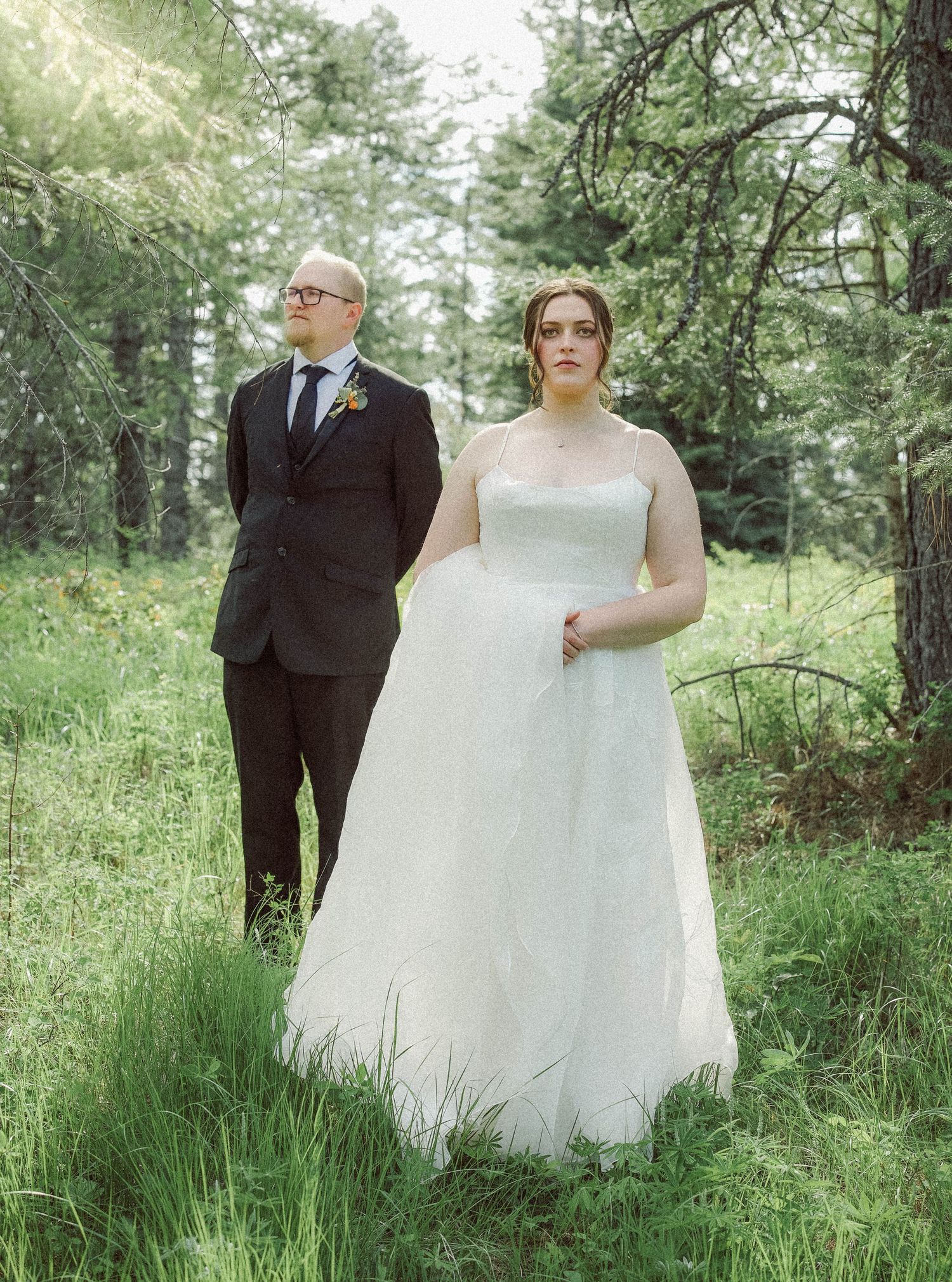 Wedding couple stands elegantly in a lush green forest, bride in flowing white gown, groom in dark suit.