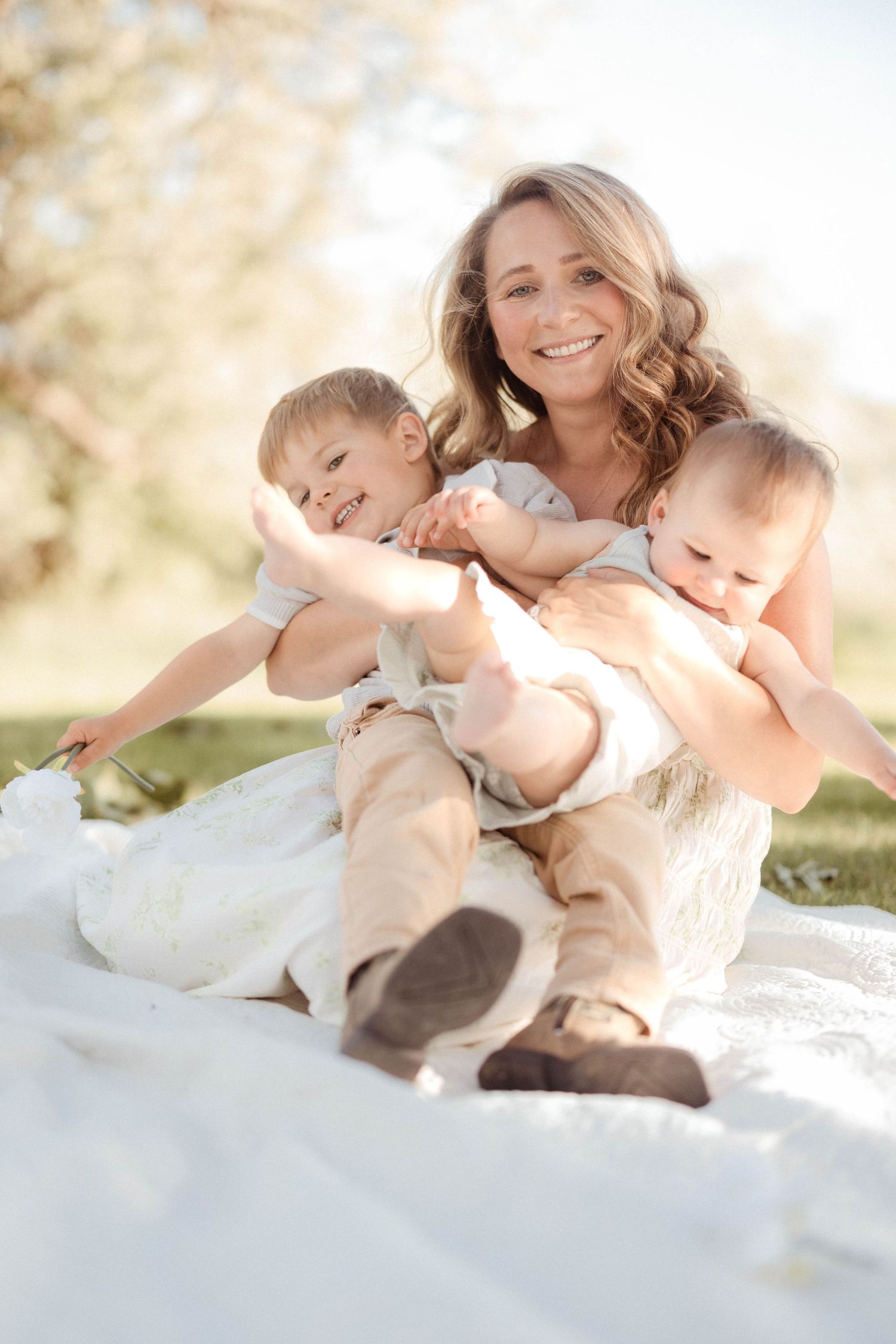 A smiling mother sits on a white blanket outdoors joyfully holding her two young children in golden sunlight.