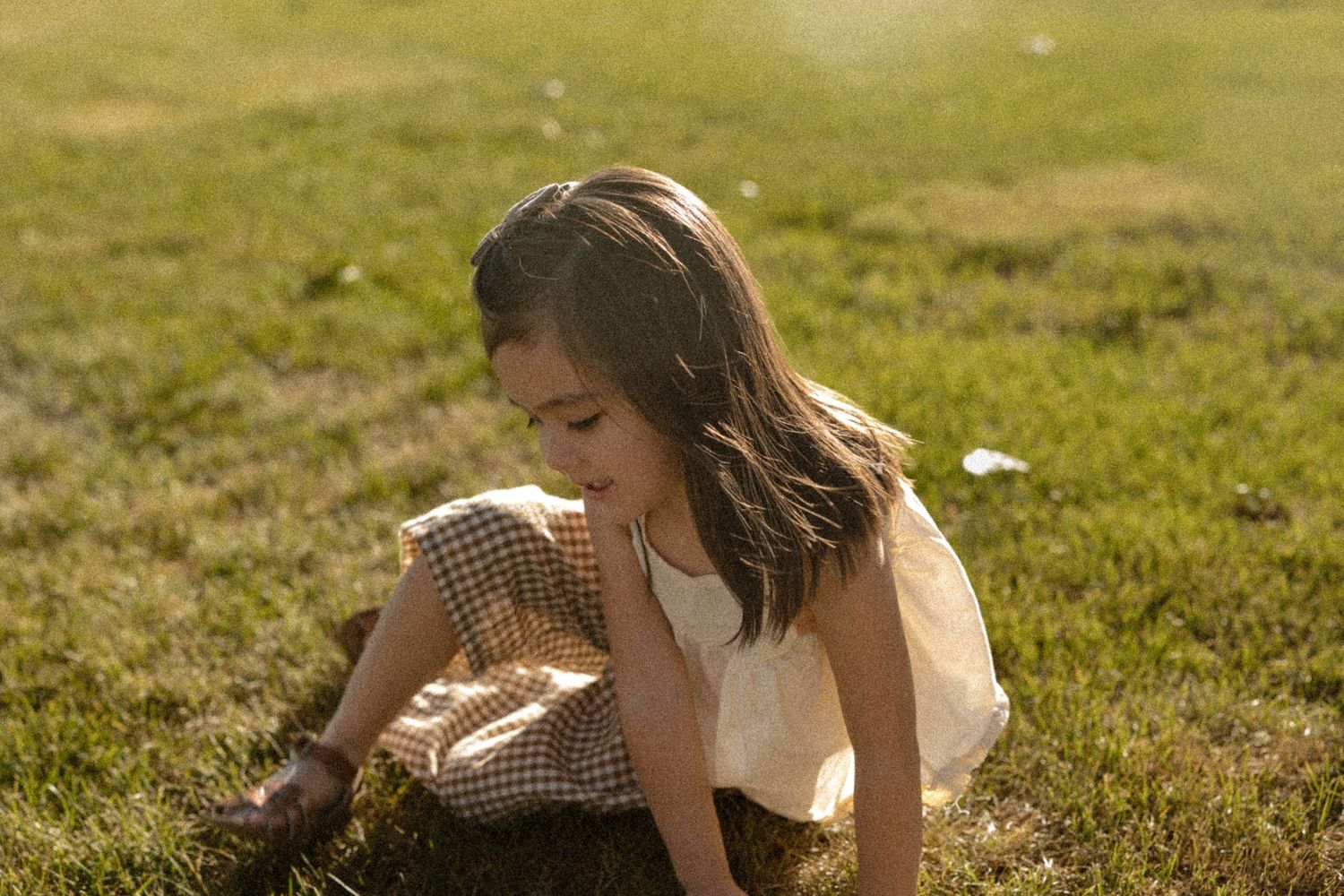 A young girl in a checkered dress plays on green grass in warm golden sunlight.