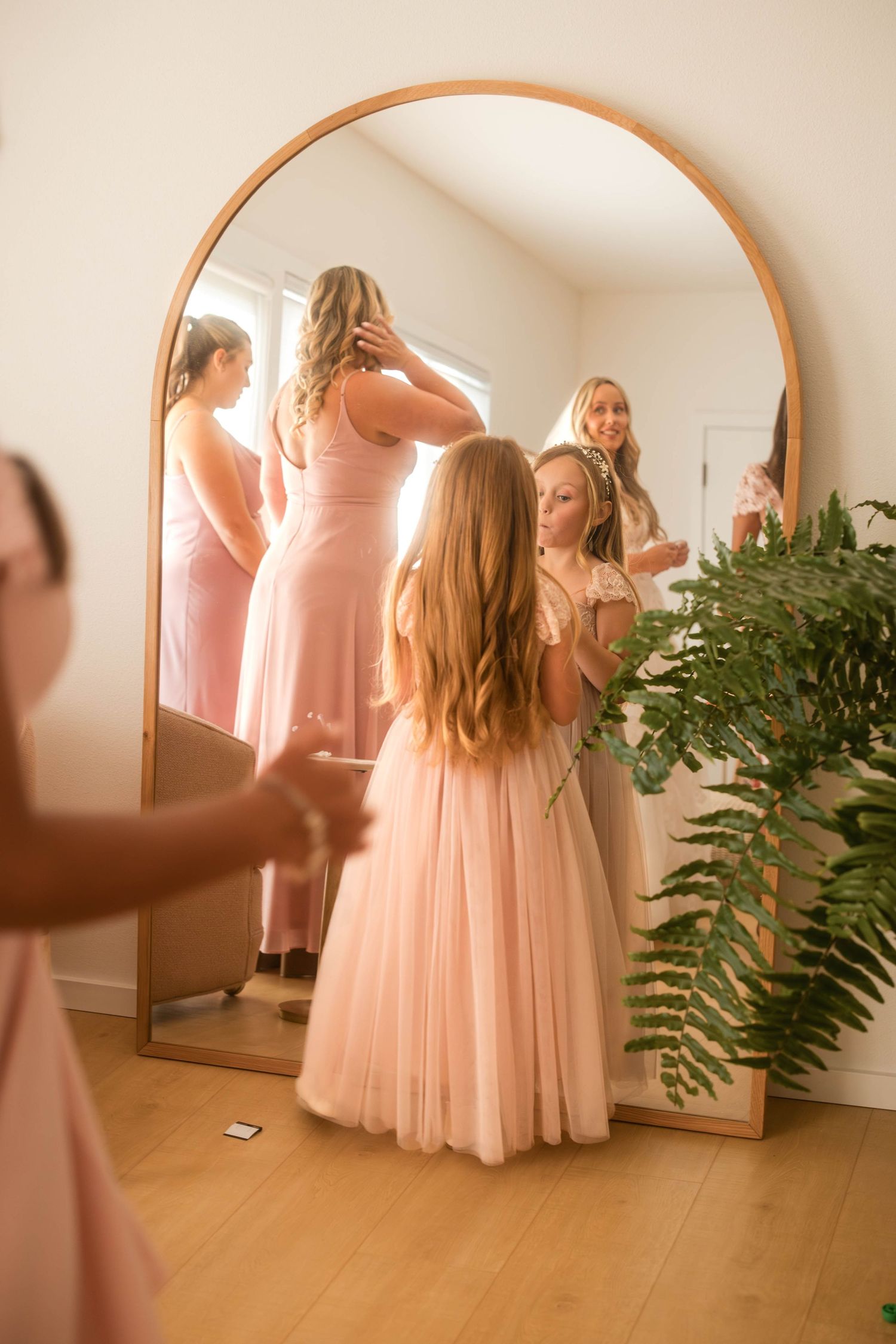 Bridesmaids in pink dresses getting ready together, reflected in a large arched gold mirror with a flower girl.
