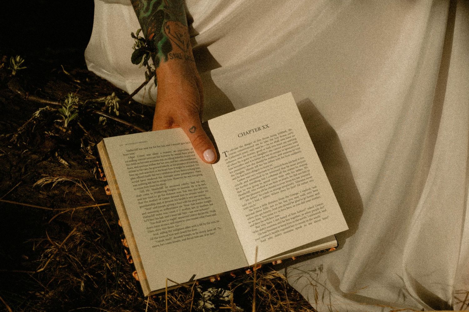 Tattooed hand holding an open book with white fabric in background, outdoor reading in natural light.
