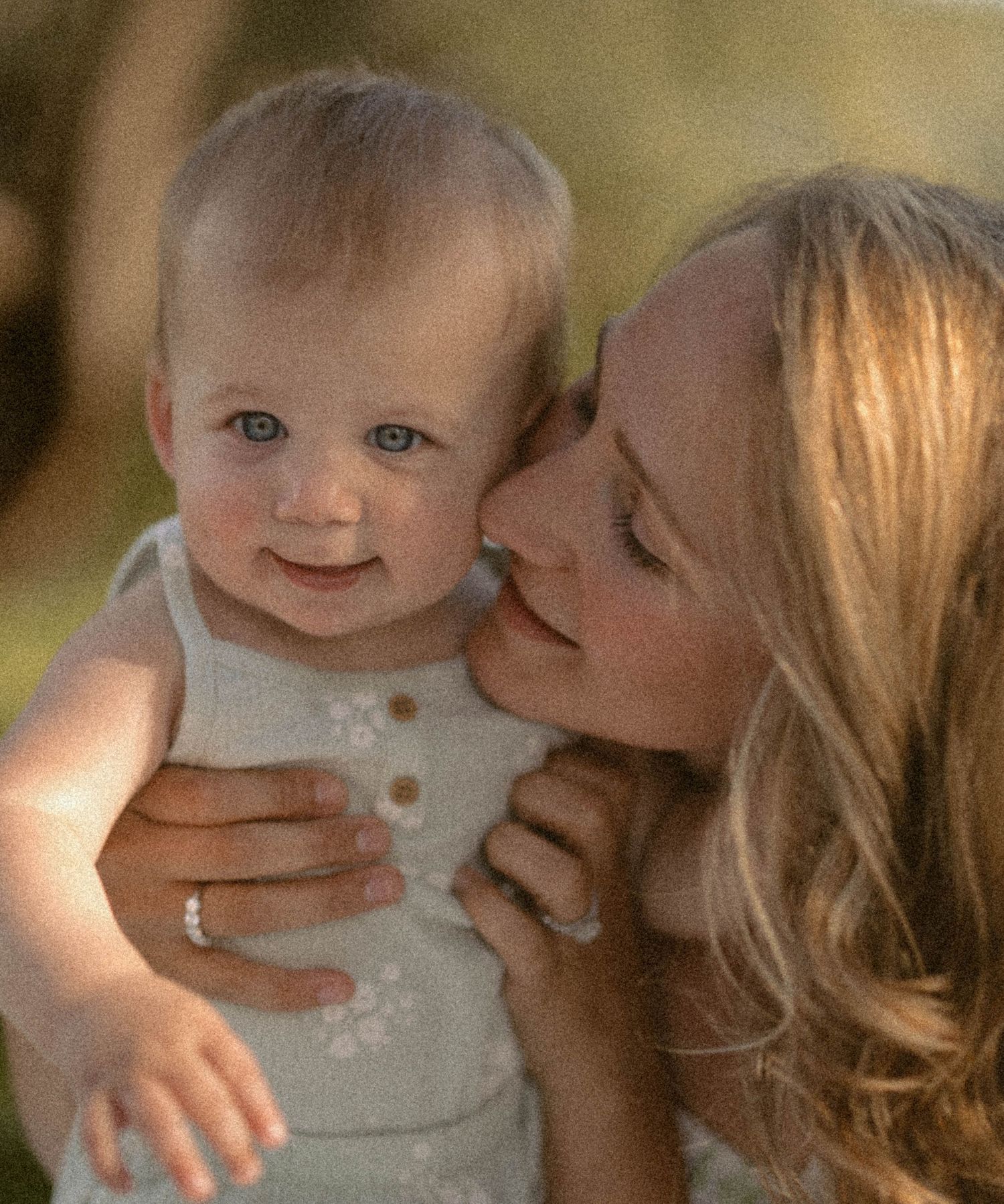 A mother tenderly kisses her blue-eyed baby outdoors in warm golden light.