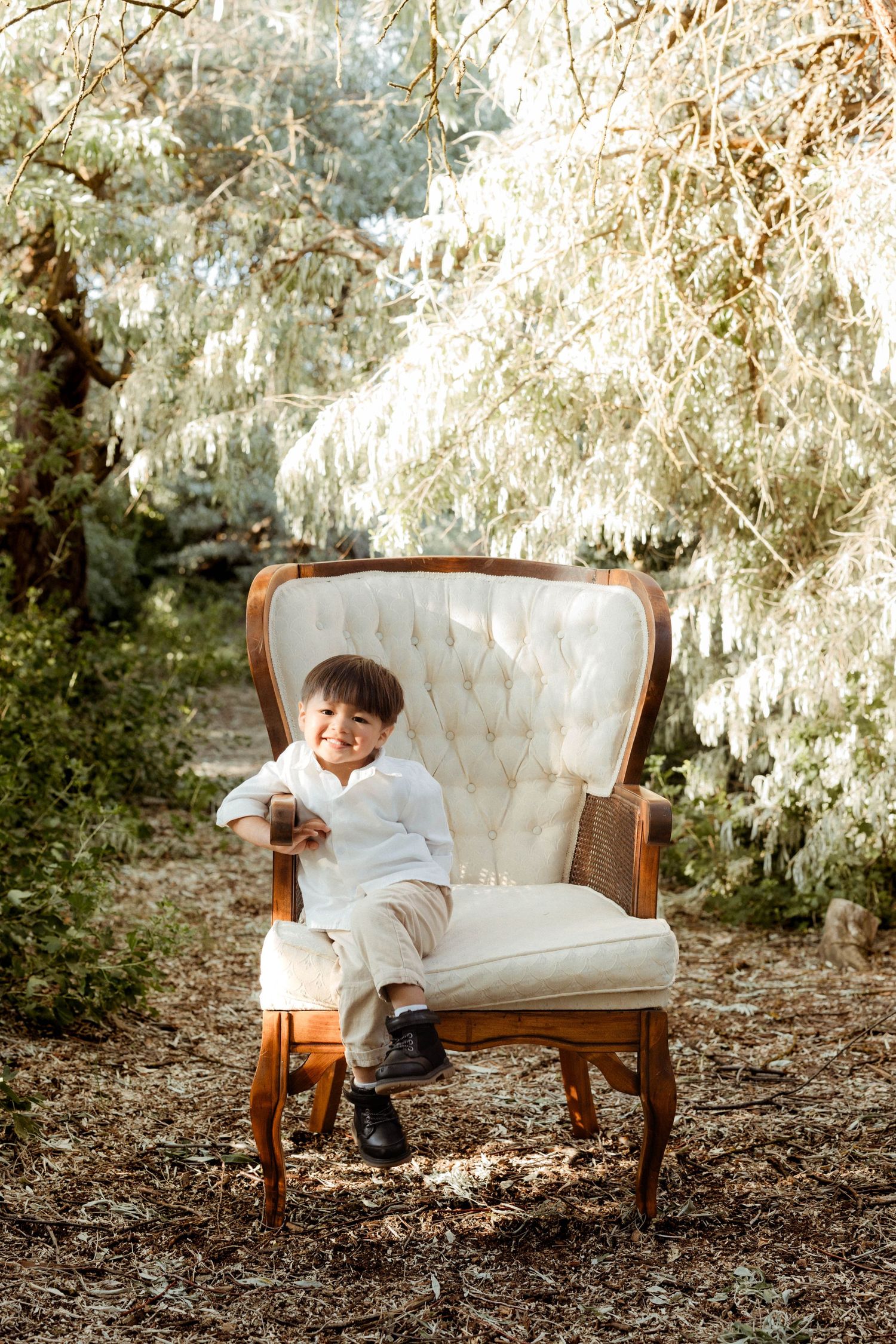 A happy toddler boy in a white shirt sits in a vintage cream armchair outdoors surrounded by white-leafed trees.