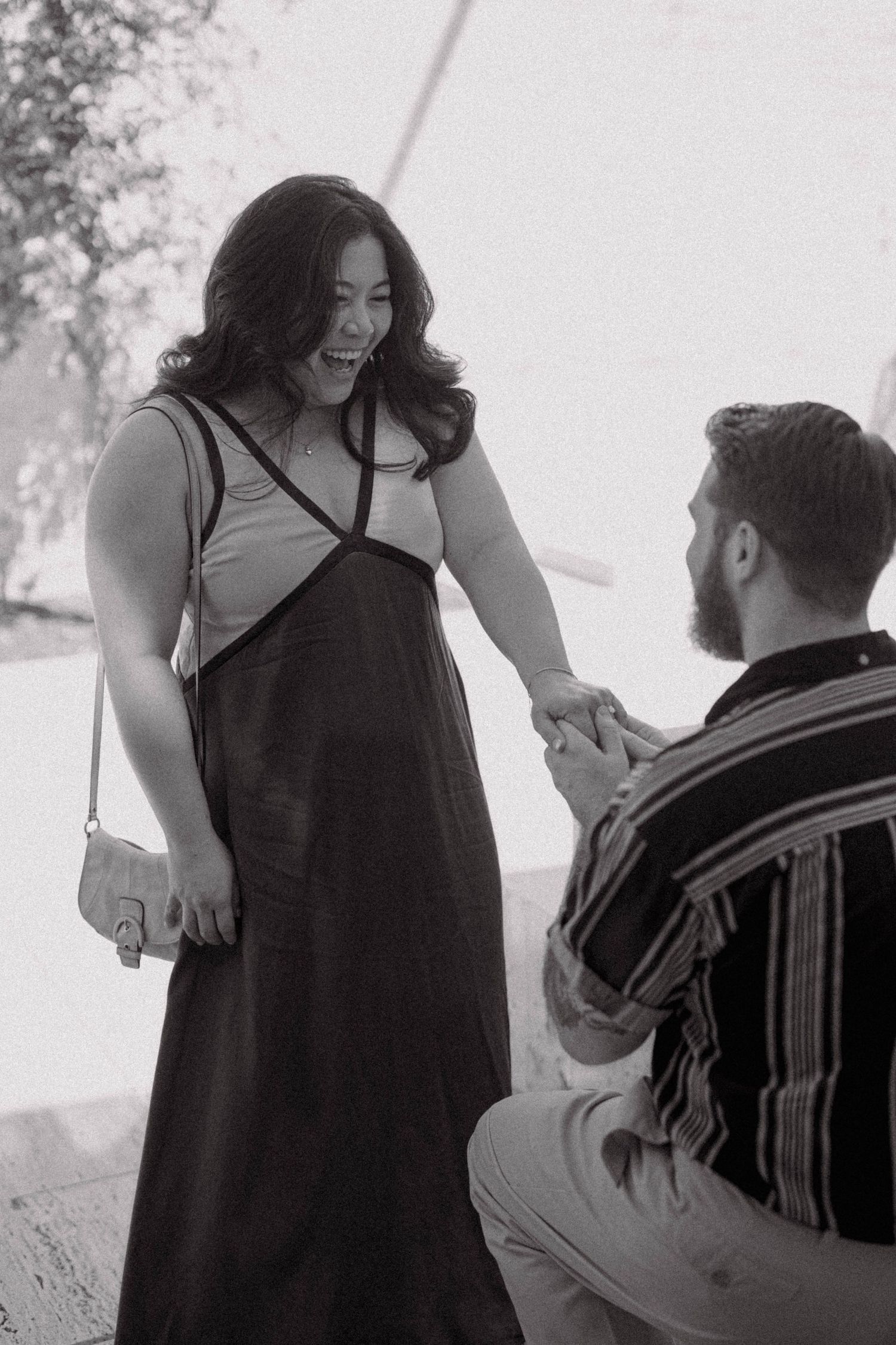 Man proposing marriage on one knee to surprised laughing woman in black and white photo.