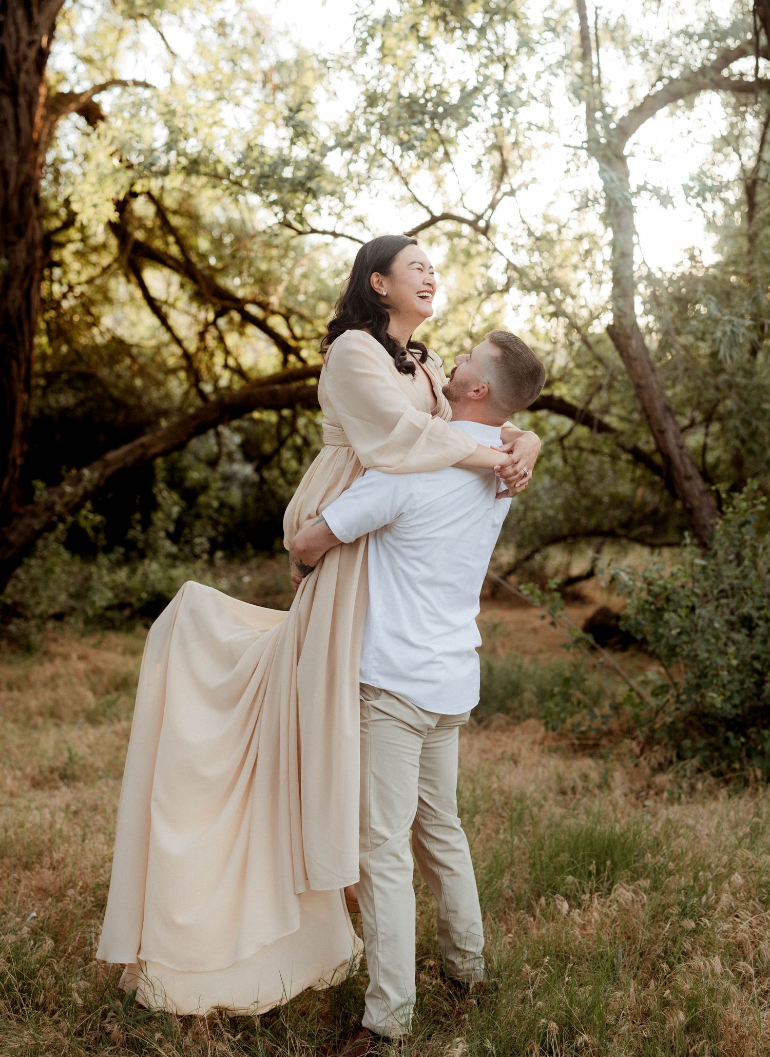 Man joyfully lifting laughing woman in beige dress during outdoor engagement session among trees.