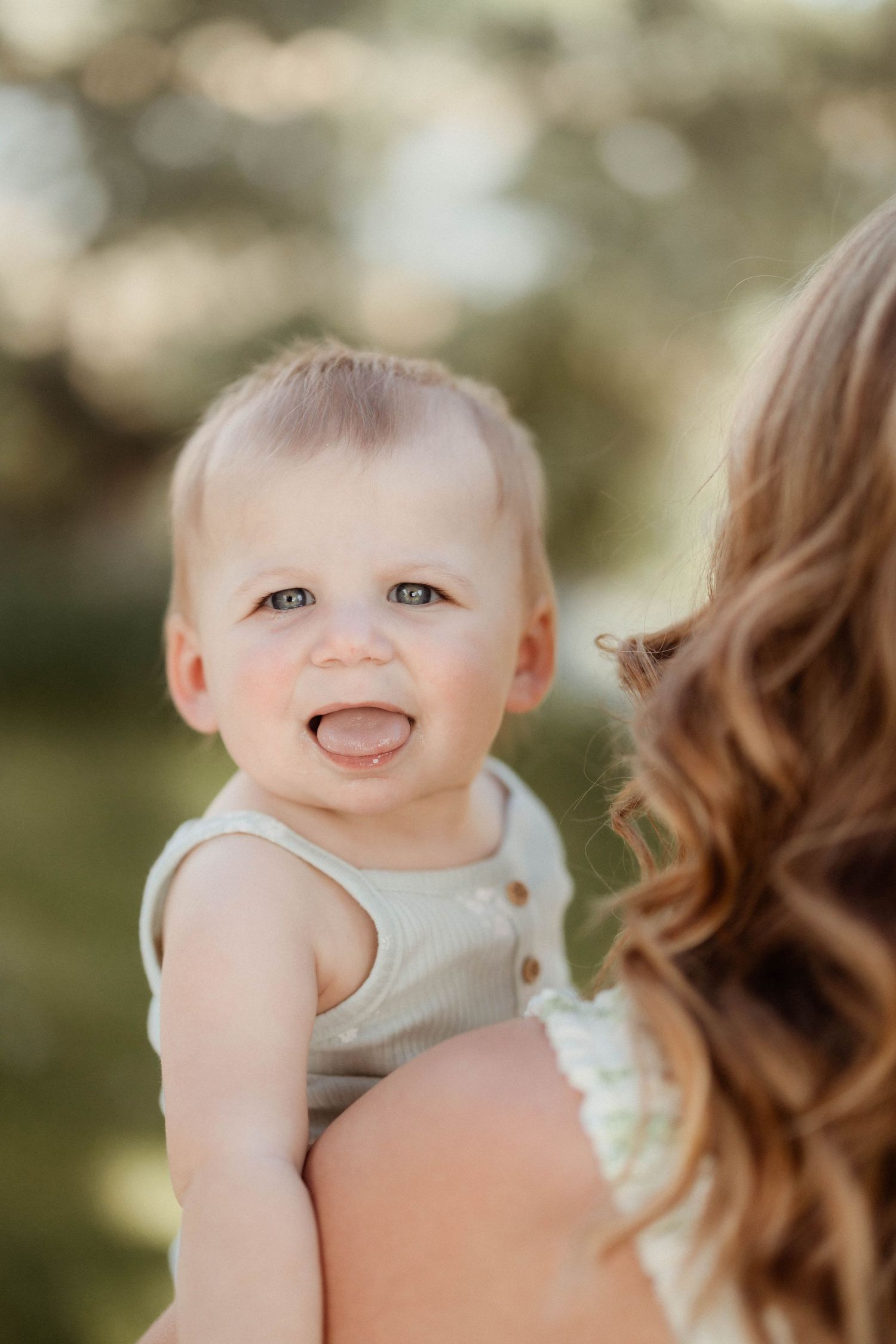 An adorable blue-eyed baby sticks out their tongue while being held by their curly-haired mother outdoors.