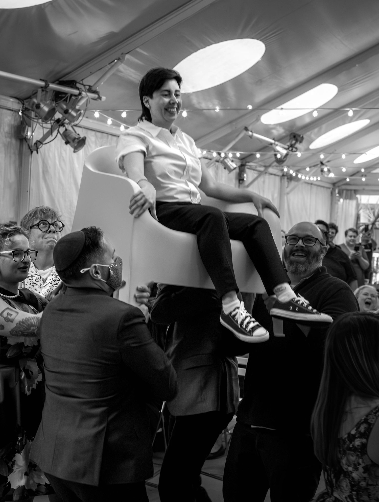 Black and white photo of a smiling guest being lifted in a chair during a wedding celebration.