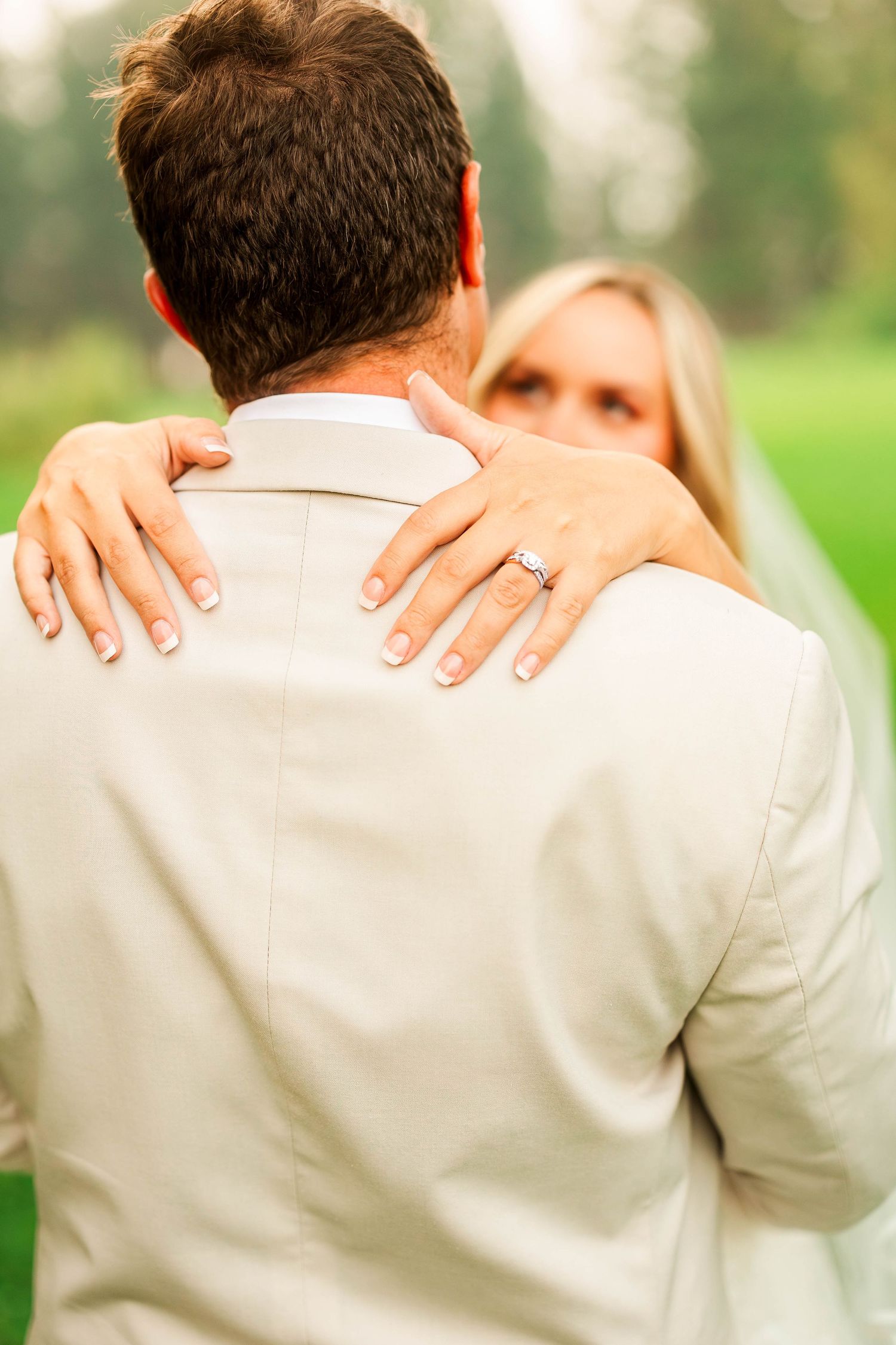 Close-up of bride with diamond ring embracing groom from behind on lush green wedding venue grounds.