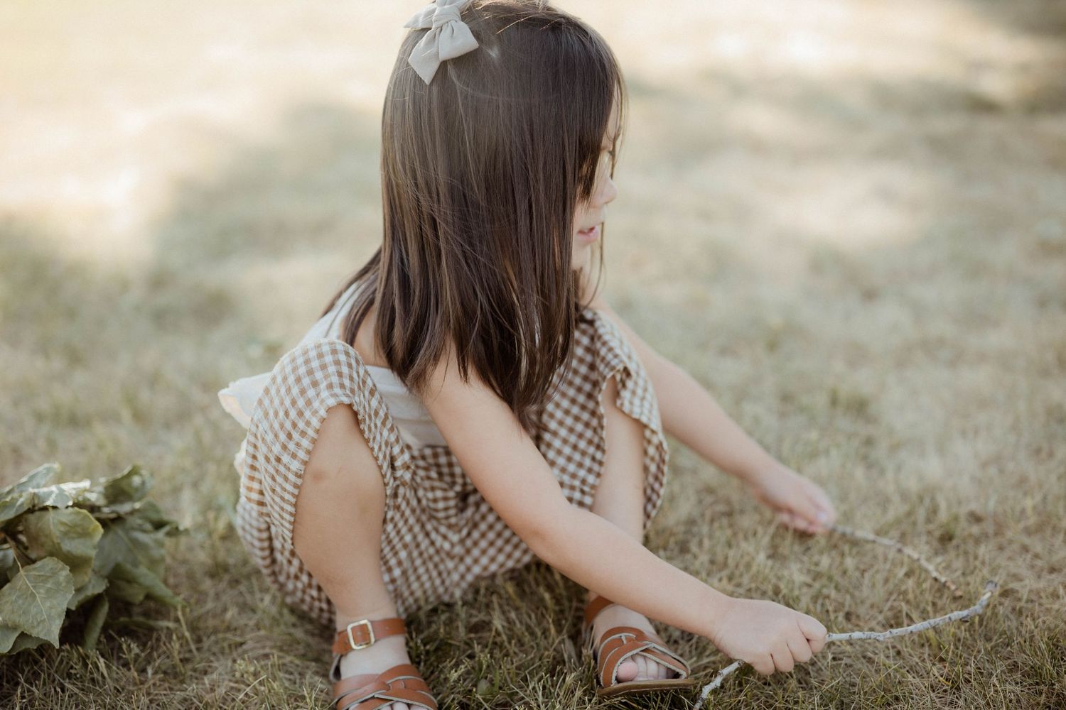 Young girl in checkered dress with hair bow sitting on grass in warm sunlight.