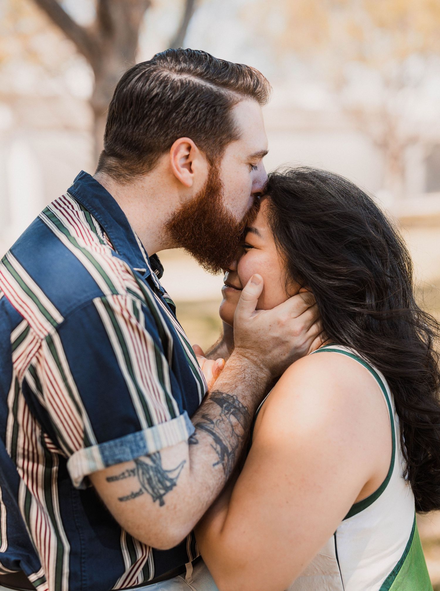 A bearded man with tattoos gently kisses a dark-haired woman outdoors among autumn trees.