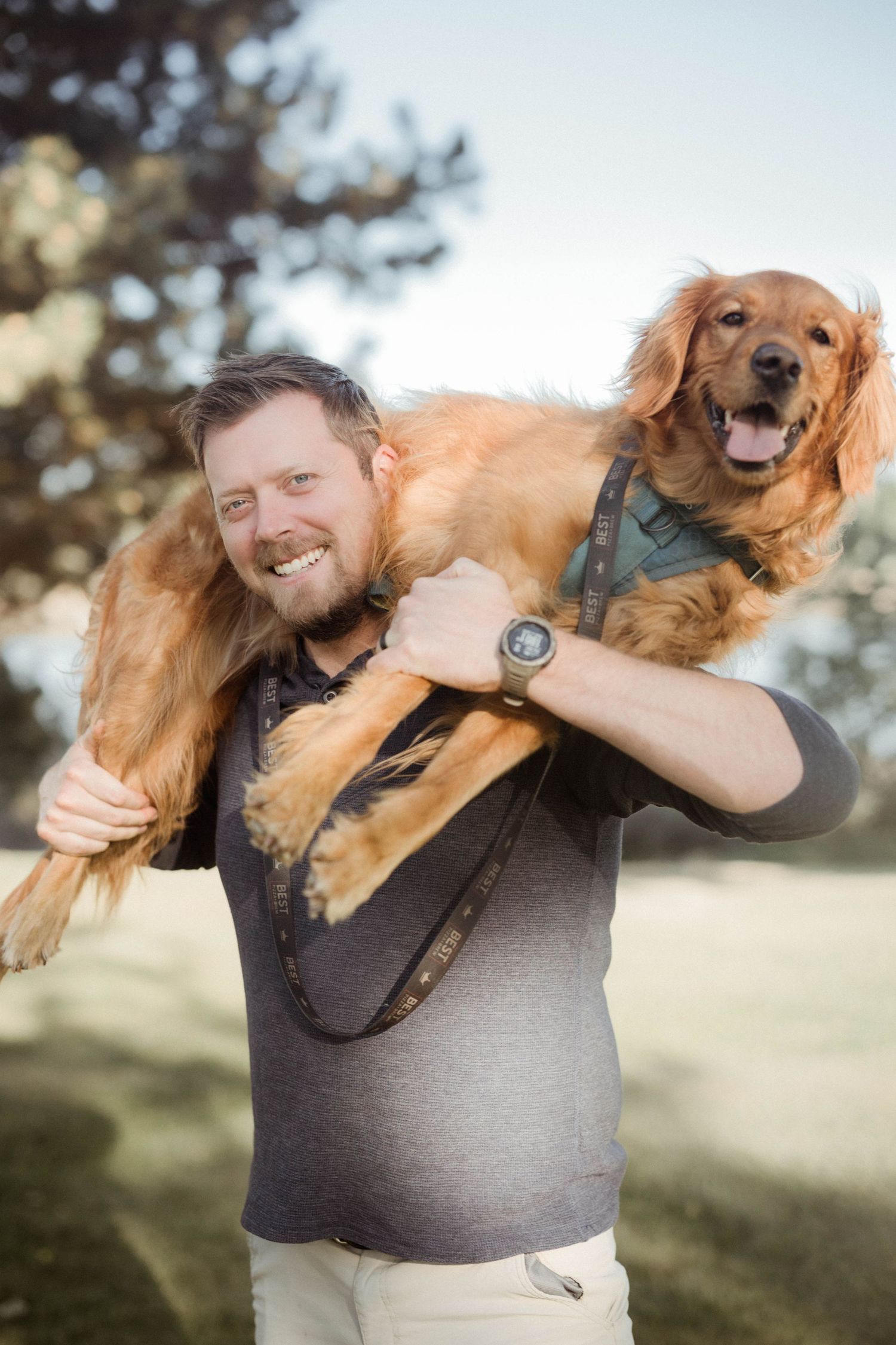 A smiling man carries a happy golden retriever on his shoulders in an outdoor park.