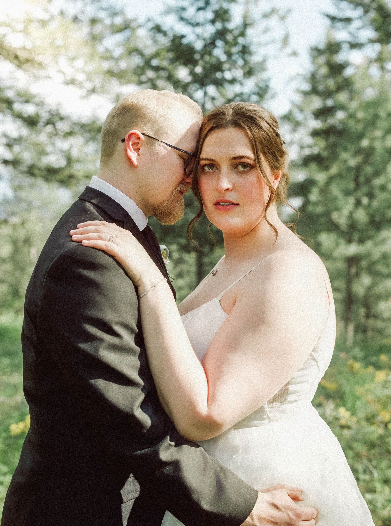 Bride and groom share an intimate embrace in a sunlit forest, bride in white gown, groom in black suit.