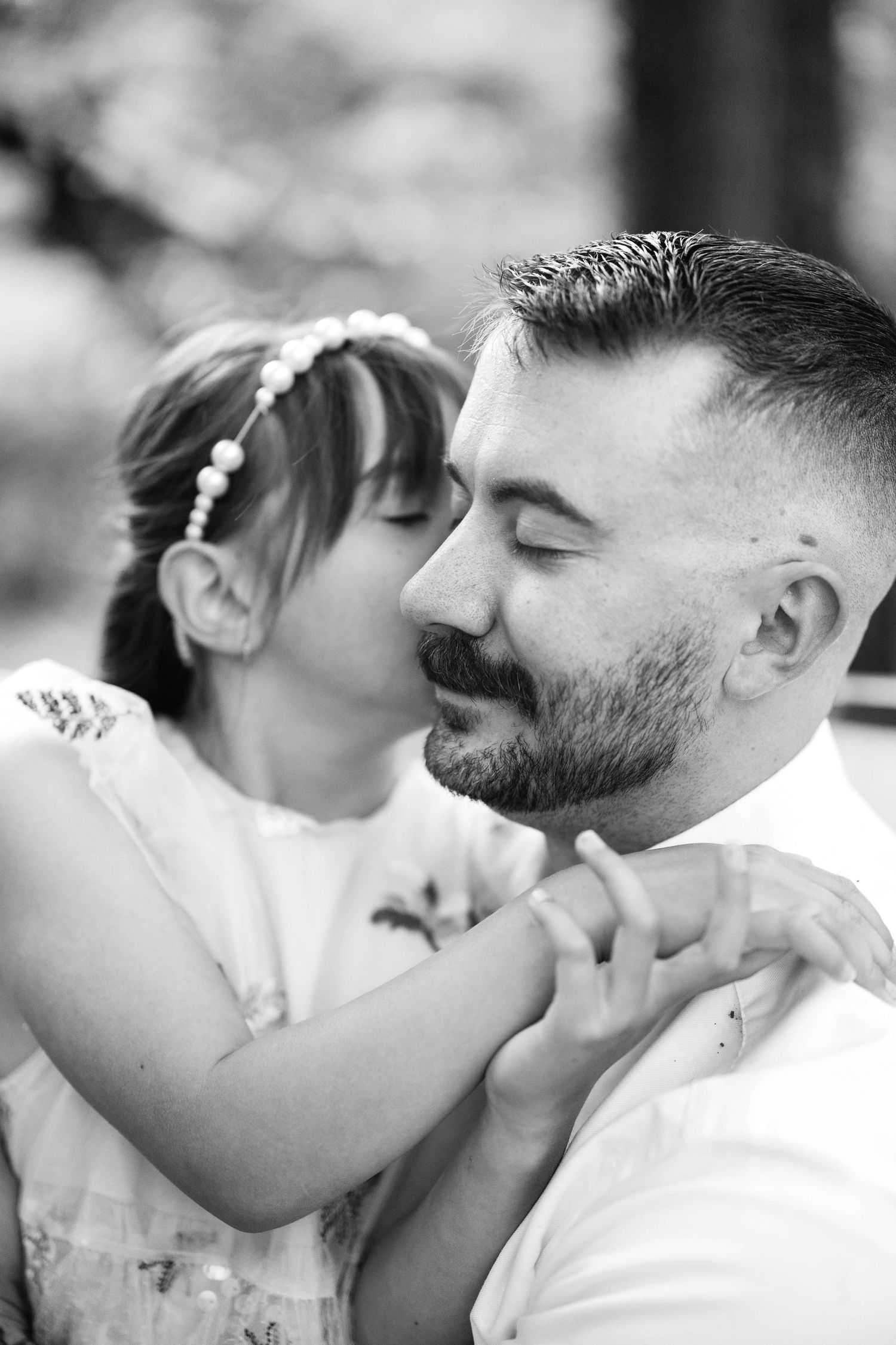 A black and white photo of a young girl kissing her bearded father on the cheek outdoors.