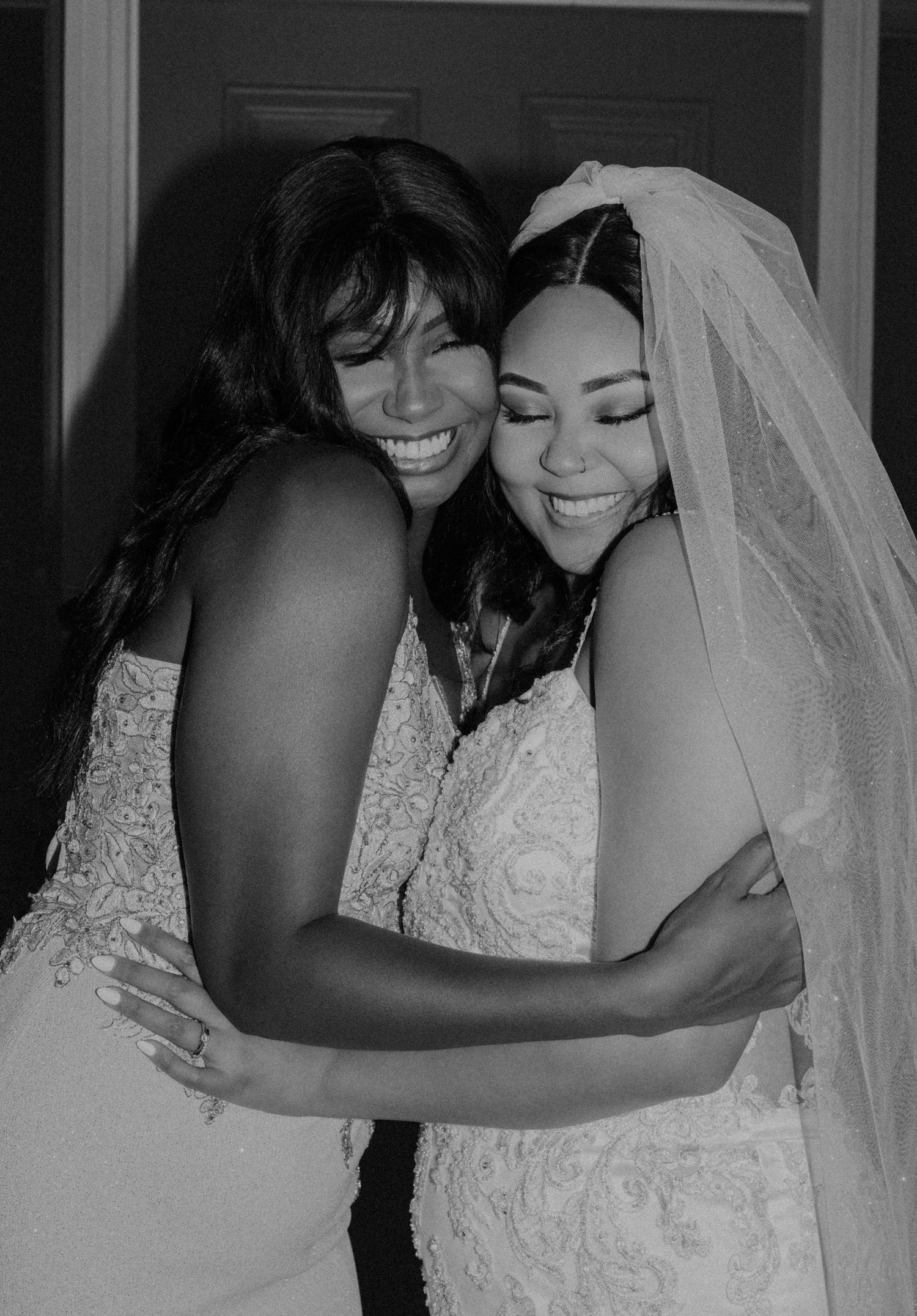 Two women in wedding gowns share a joyful embrace in a heartfelt black and white wedding photo.