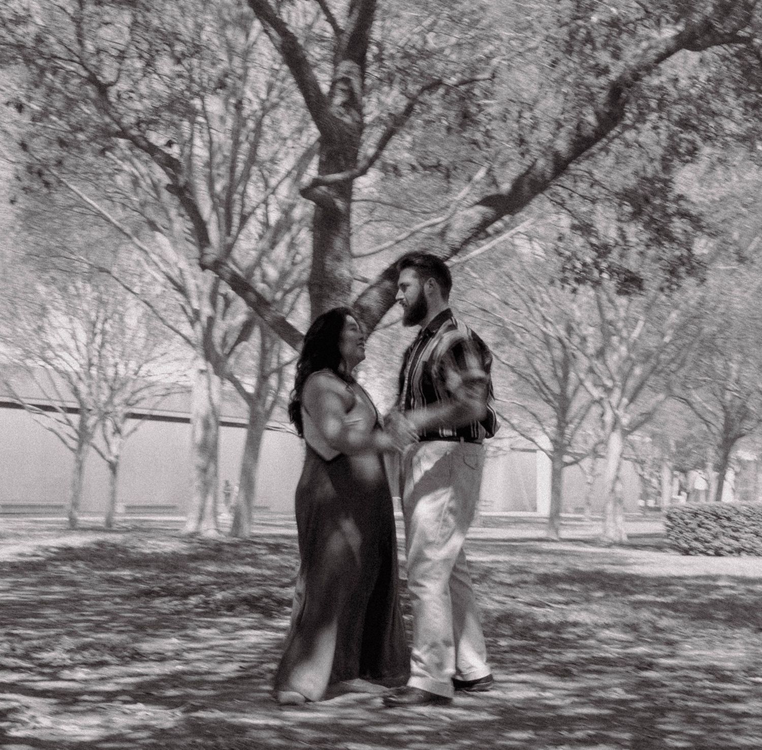 Black and white photo of a couple dancing together among trees in an outdoor park setting.