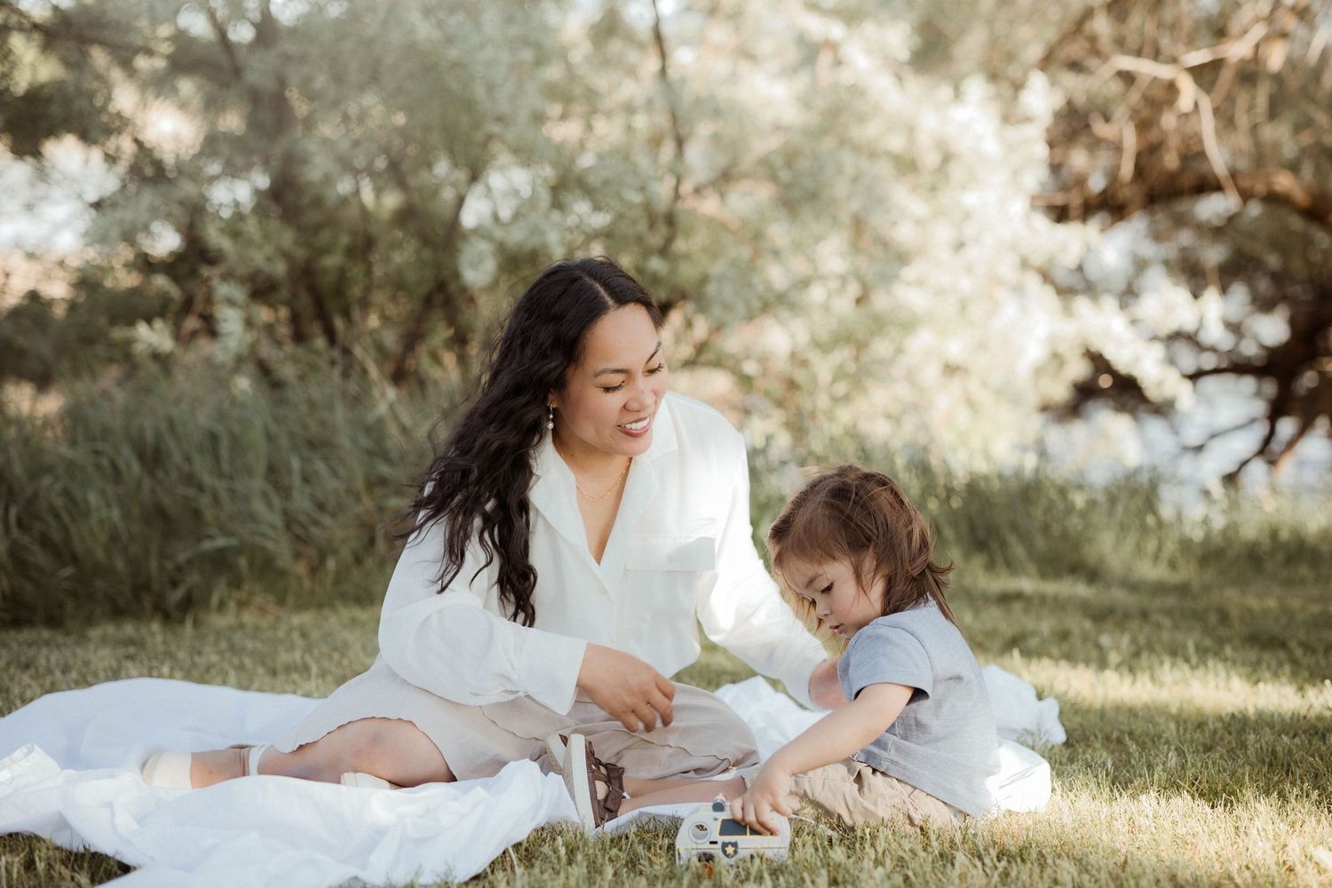 Mother in white outfit sitting on blanket with toddler child playing in green park.