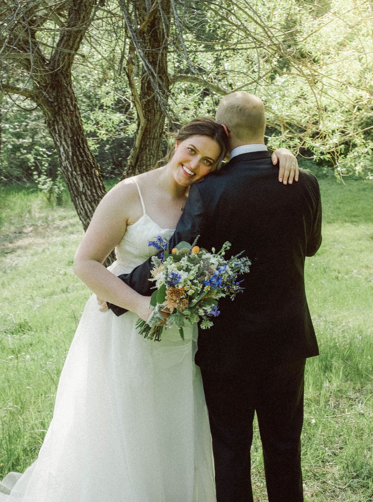 Smiling bride in white gown hugging groom while holding a colorful wildflower bouquet outdoors.