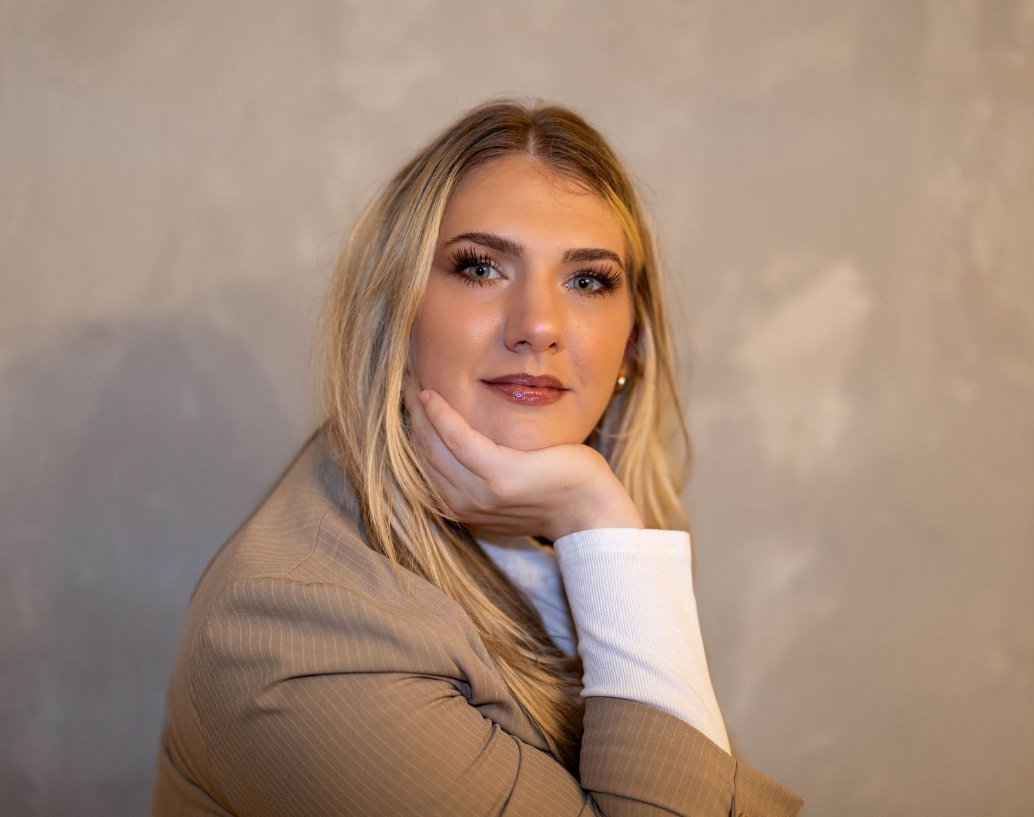 Elegant blonde woman posing confidently against a textured gray wall in a beige blazer.