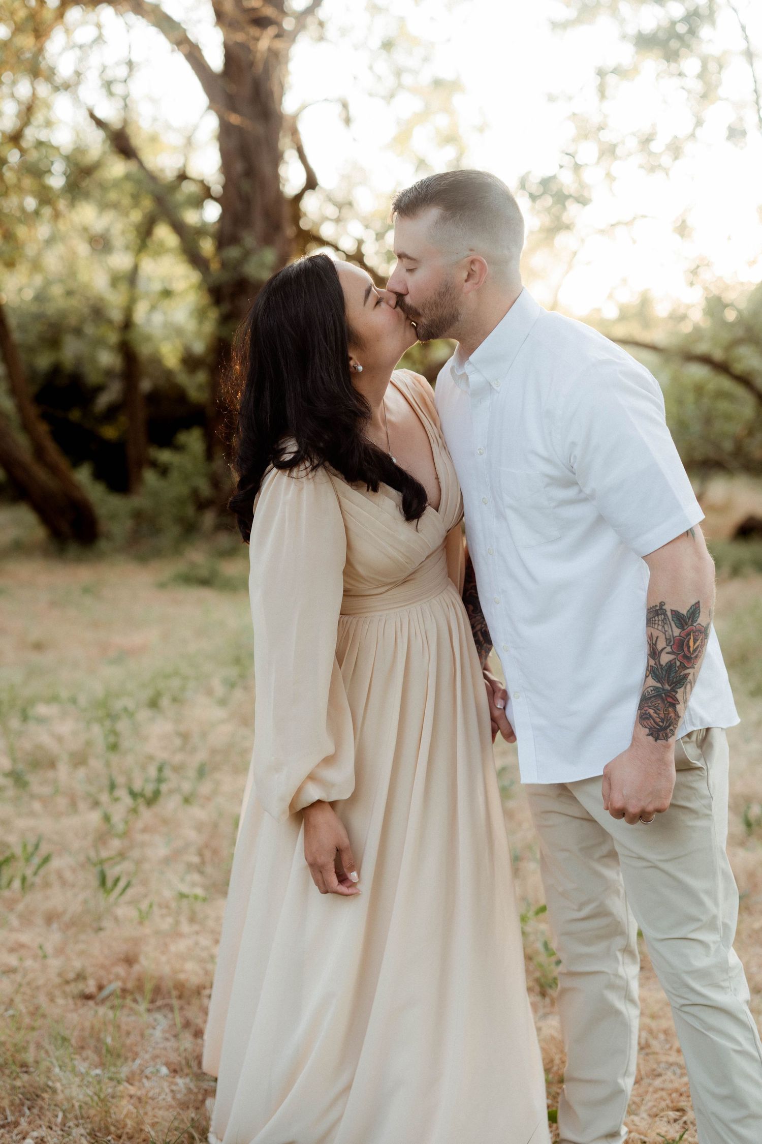 Couple sharing a tender kiss outdoors, woman wearing elegant beige flowy dress, man with tattoo sleeve.