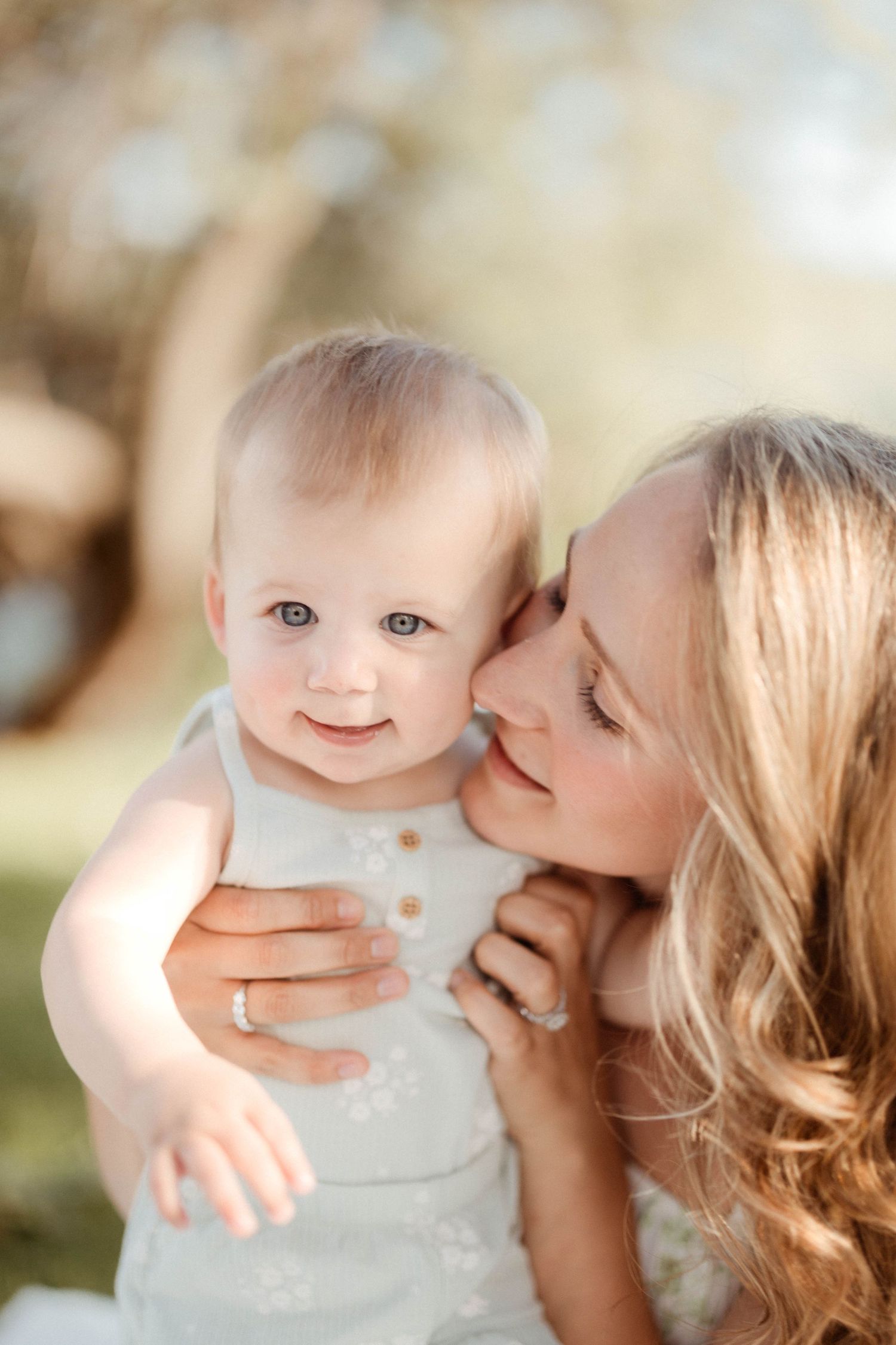 A loving mother with curly hair kisses her blue-eyed baby girl on the cheek in a sunny outdoor setting.
