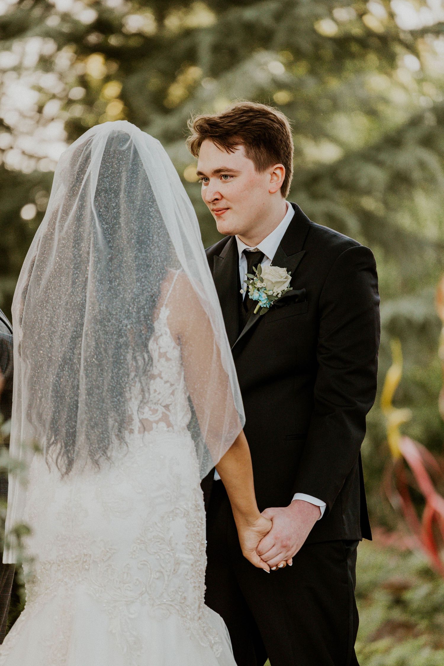 A groom in a black tuxedo smiles at his veiled bride during an outdoor wedding ceremony.