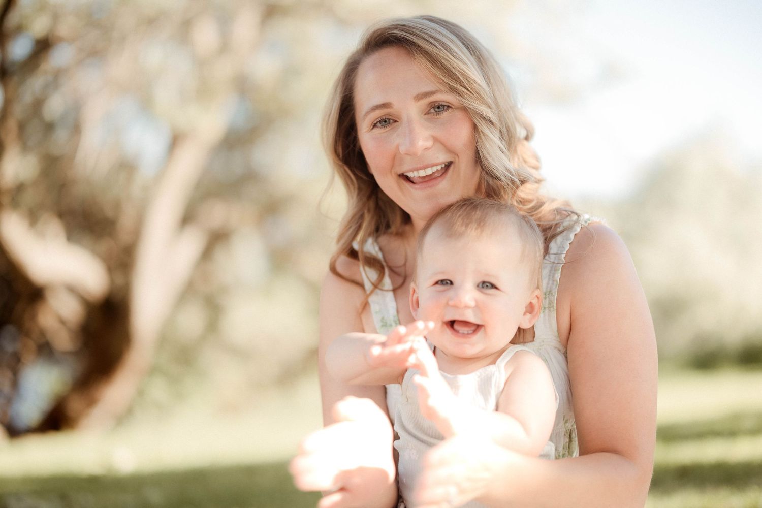 A happy mother and her laughing baby pose together in a bright sunny outdoor setting.