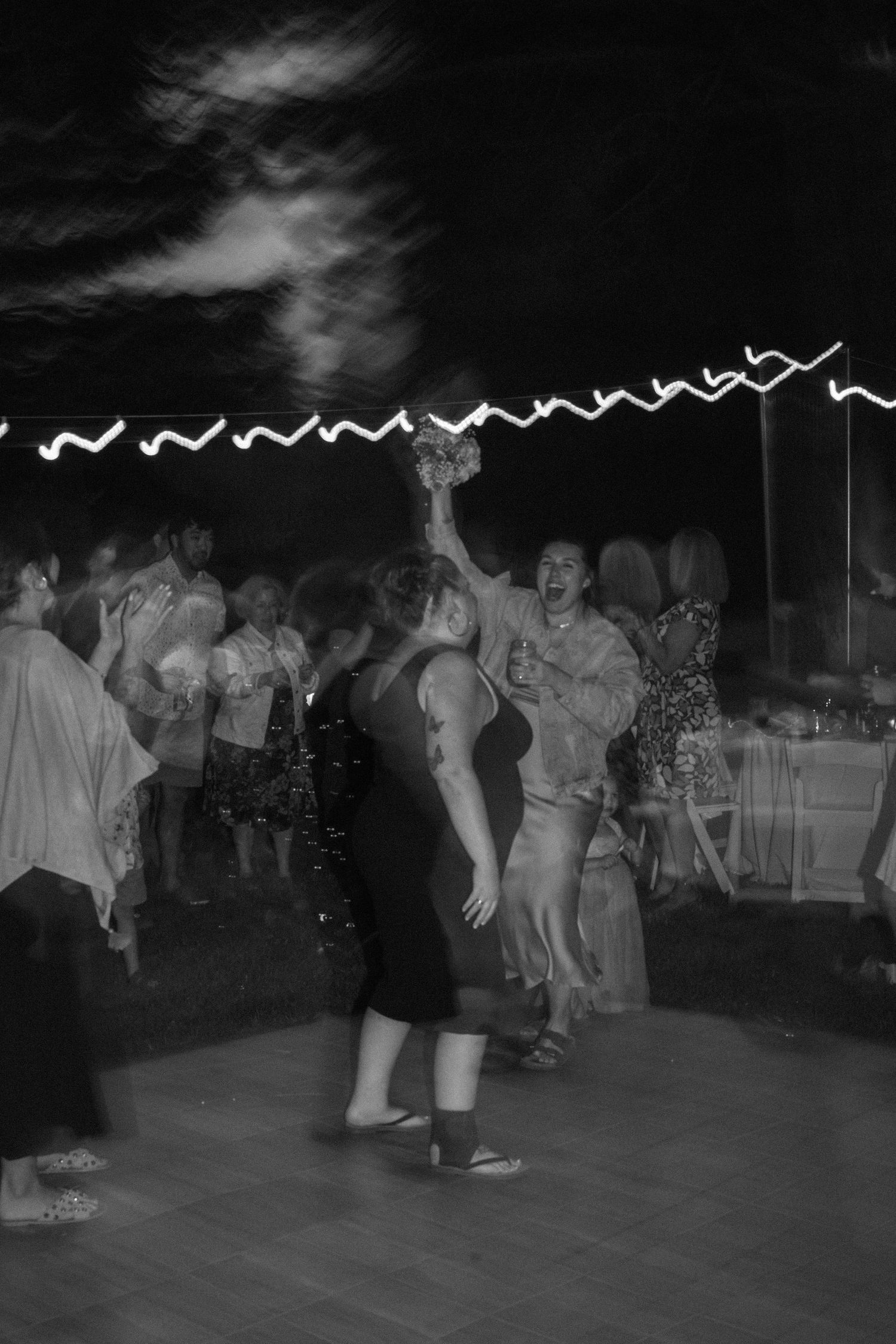 Black and white photo of guests dancing and catching bouquet at lively outdoor wedding reception at night.