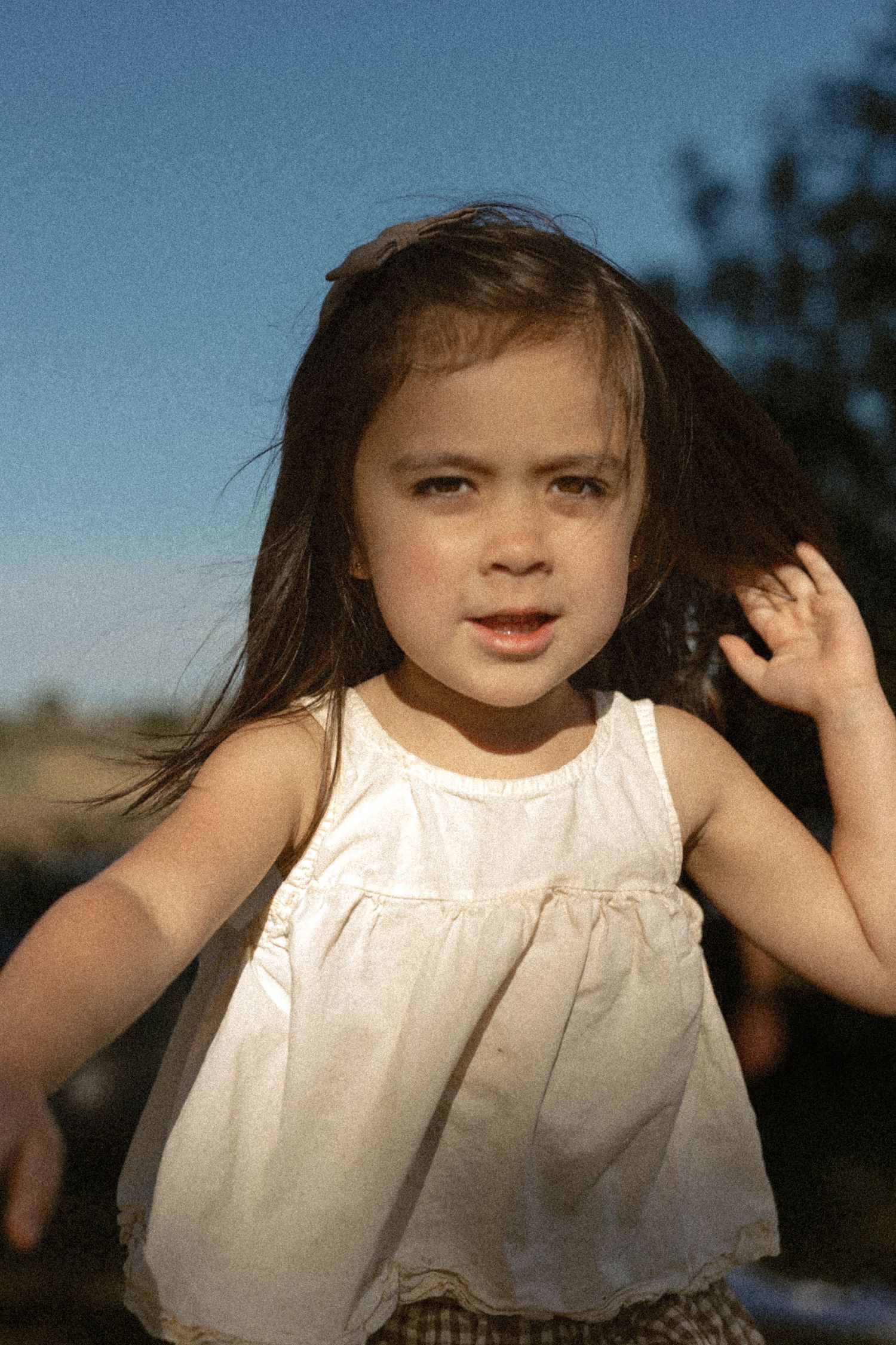 A young girl in a white sleeveless top with windswept dark hair looks at the camera outdoors.