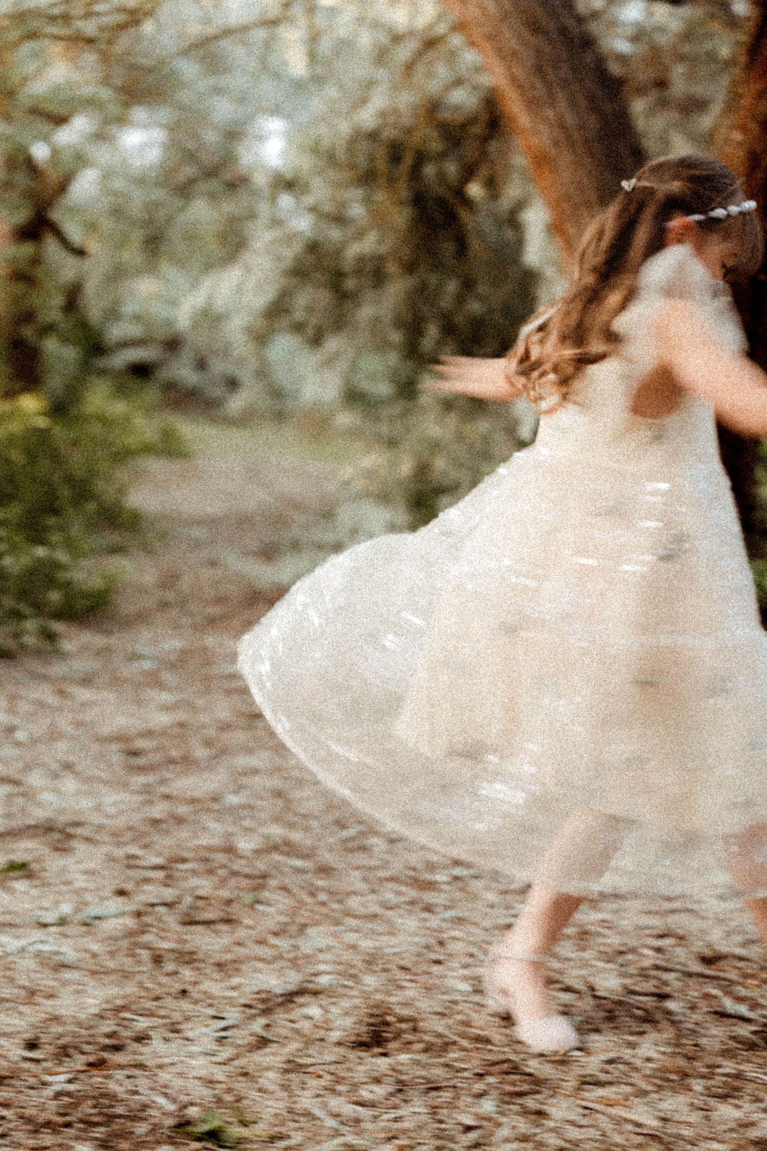 A young girl in a white tulle dress twirls barefoot on a forest path, captured in motion blur.